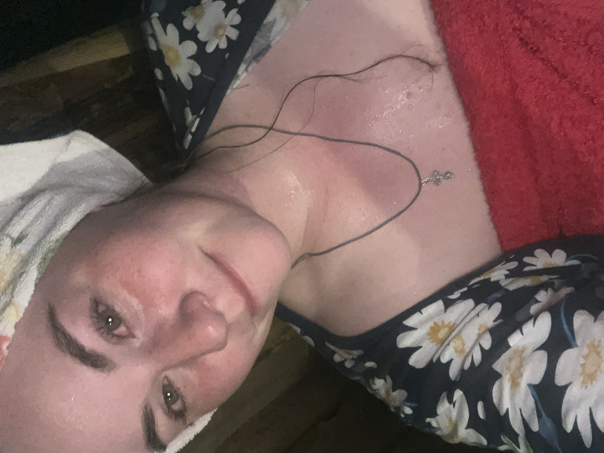 A selfie shot of a fair-skinned person with a white towel wrapped around their head. The person has a floral print top on. A dark cord necklace with a cross pendant is visible on their chest. The person appears sweaty. A red towel is positioned to the right. The setting appears to be indoors on top of wood. The image was taken in Сосново, Russia.