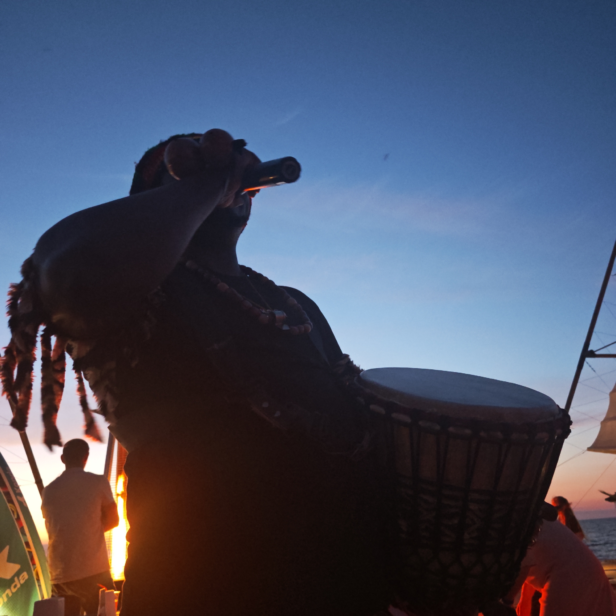 This is a low-angle, silhouette shot of a person playing a djembe drum against a twilight sky. The person has dreadlocks and is wearing beads. In the background, a person in a light-colored shirt is visible, standing near some tall structures that appear to be part of a stage or scaffolding. There is a fire or bright light source to the left, illuminating the scene with an orange glow. The location is described as a municipal settlement in Shelkino, Ukraine, with the sea visible in the distance. The overall atmosphere is one of a lively outdoor performance at dusk.