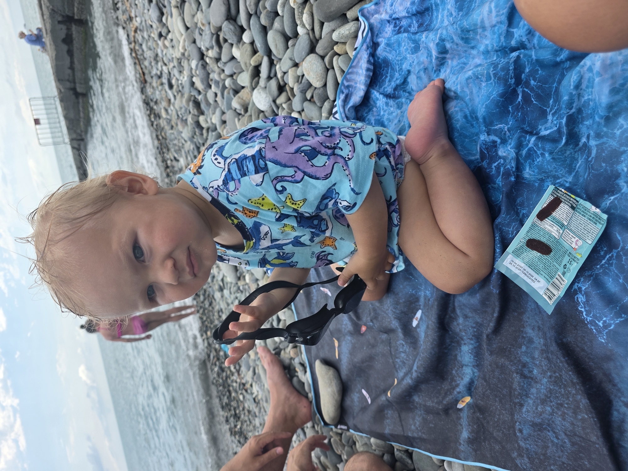 A young child with blonde hair and blue eyes sits on a blue towel with a wave pattern on a pebble beach. The child is wearing a light blue t-shirt with an octopus and sea creature print. The child is holding a pair of black swimming goggles in their right hand. A snack wrapper is lying on the towel in front of the child. In the background, the sea is visible with waves breaking on the shore. Other people are also present on the beach and in the water, including a woman in a pink bikini and a person in a blue shirt sitting on a concrete pier. The sky is overcast. The location is identified as Sochi, Russia.