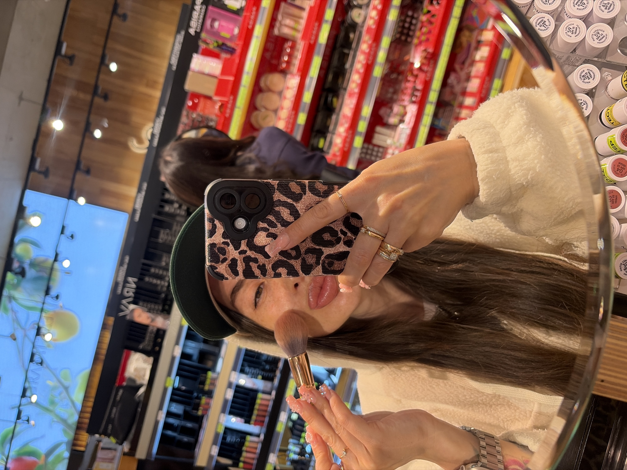 A woman with long dark hair, wearing a green baseball cap and a cream-colored fuzzy jacket, is taking a mirror selfie in what appears to be a cosmetics store. She is holding a makeup brush to her cheek and her phone, with a leopard-print case, is positioned to capture the reflection. Behind her, shelves filled with makeup products are visible, with red and green shelving units. The woman is wearing multiple rings on her fingers and a wristwatch. The background suggests an indoor retail environment, possibly a department store or a specialized beauty shop. The reflection in the mirror also shows other shoppers and the store's lighting fixtures.