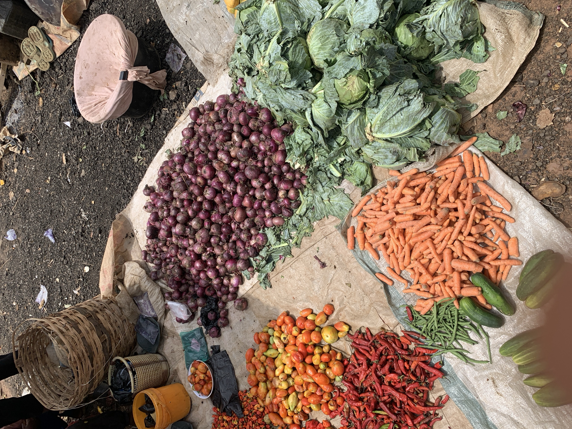 A high-angle shot reveals a diverse assortment of produce spread across what appears to be a beige tarp, likely in an outdoor market setting in Keffi, Nigeria. Predominantly featured are large piles of red onions, heads of green cabbage, a heap of orange carrots, tomatoes in various shades of red and yellow, and a collection of red chili peppers. Interspersed are cucumbers and green beans.
To the left, a woven basket sits near an assortment of smaller containers, some with visible contents. A wheel covered in fabric is in the upper-left corner of the frame. The ground surrounding the tarp is primarily brown and rocky. No individuals are visible in the immediate frame.