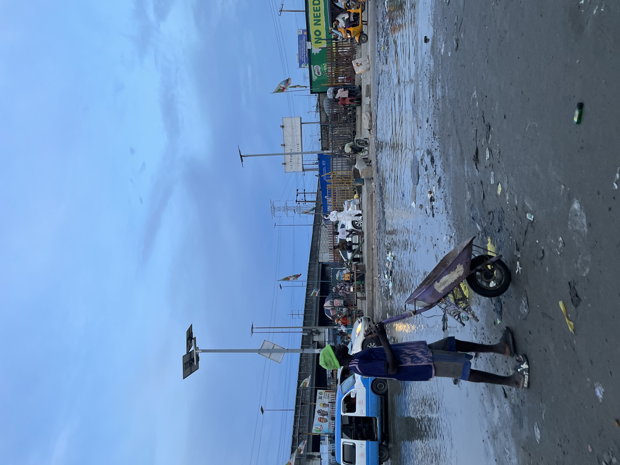 A young boy wearing a green hat and a blue and purple patterned shirt is pushing a purple wheelbarrow across a flooded street in Lekki, Nigeria. The street is filled with debris, including trash and plastic bottles. In the background, there is a blue and white van, a gold car, a yellow tricycle, and several people walking and standing around. There are also a few flags and billboards visible. The sky is overcast, and the overall impression is one of a busy, somewhat unkempt urban environment.