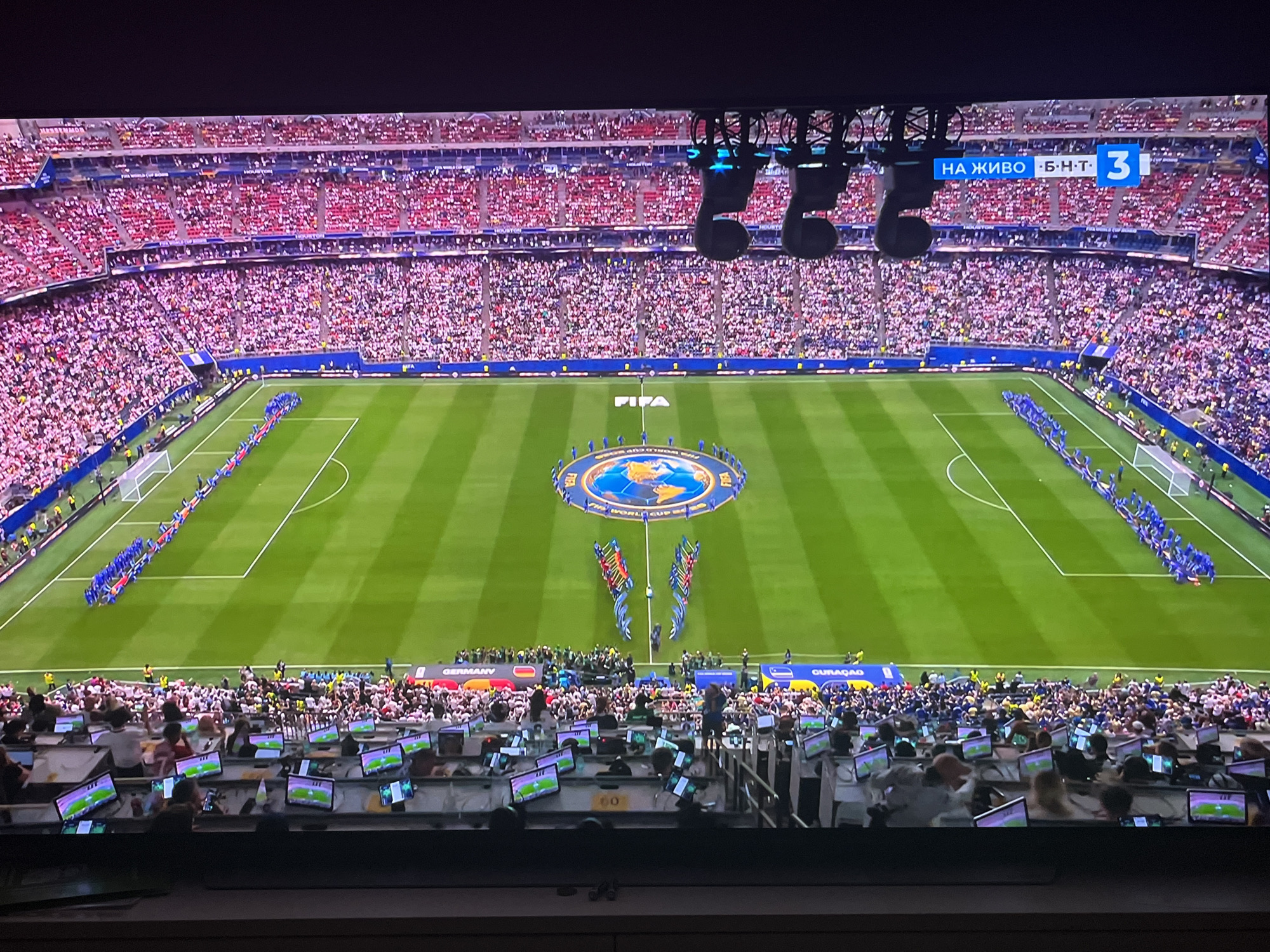 The image shows a wide shot of a soccer stadium filled with spectators. In the foreground, a large number of people are seated at desks, each with a monitor in front of them, appearing to be working in a press or media area. They are looking at their screens, and some are interacting with their equipment.
The middle ground of the image shows a soccer field with a large circular emblem at its center. Players are lined up on either side of the field, facing each other, possibly for a pre-game ceremony or announcement. Some officials or staff are visible on the field.
The background is dominated by stadium seating, densely packed with a large crowd of spectators dressed in various colors, predominantly red and white. Above the stadium seating, part of the city of Varna, Bulgaria is visible, with buildings and urban infrastructure. There are also large broadcast lights hanging above the field. A digital scoreboard in the upper right corner displays "НА ЖИВО · Б·Н·Т · 3".