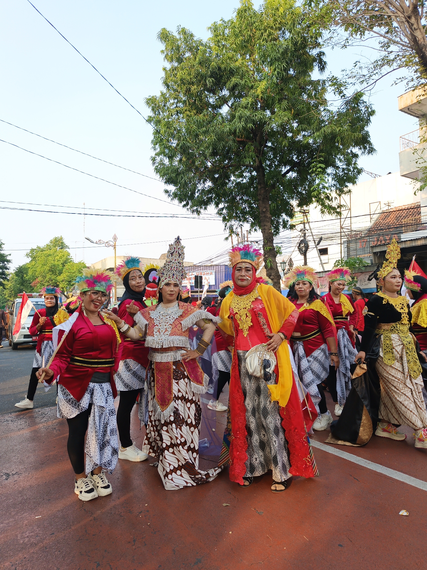 In Klaten, Indonesia, a group of women in vibrant, traditional attire participate in a parade. The women, adorned with elaborate headdresses featuring colorful feathers, wear red tops and patterned skirts. Some carry wooden sticks, while others have decorative sashes and bags. The street is paved with red asphalt, and a white line marks a boundary. In the background, trees, buildings, and a white van are visible, suggesting an urban setting. The sky is clear and pale.