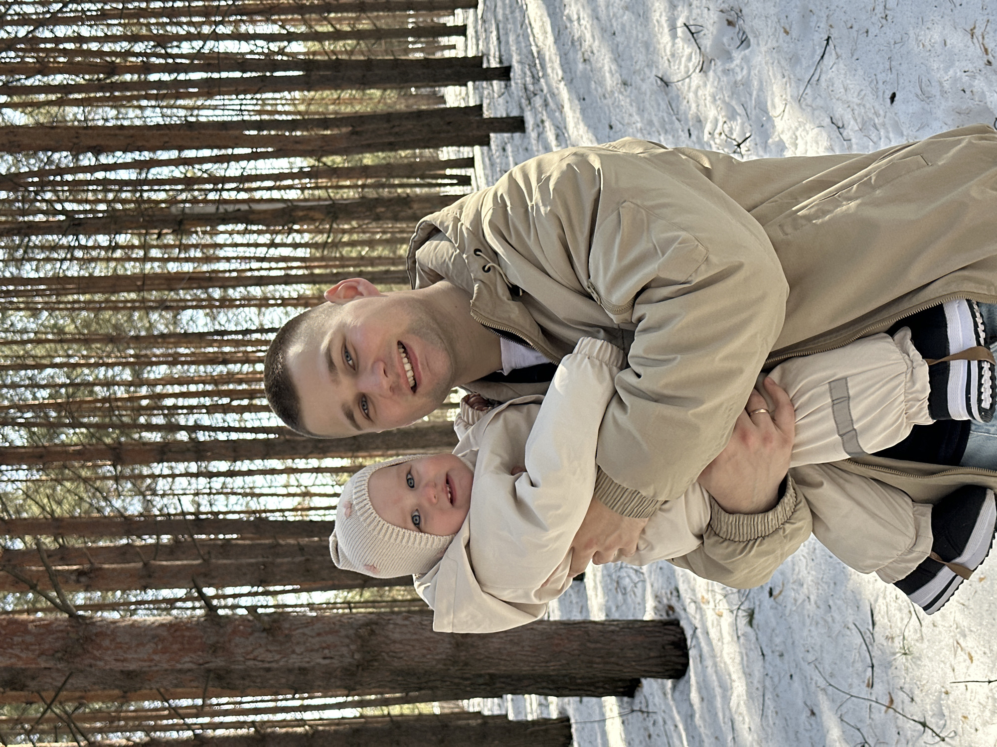 The image displays an adult male holding an infant in a snowy pine forest, likely located within or on the periphery of Chaikovsky, Russia. The ground is covered with snow, interspersed with visible patches of dark earth and dry vegetation. The adult male, appearing to be in his late 20s or early 30s, has short dark hair and light eyes. He wears a light tan or beige puffer jacket over a white t-shirt and blue jeans, with a ring visible on the ring finger of his left hand. He holds an infant, estimated to be between 6 and 18 months old, dressed in a cream-colored hooded snowsuit featuring reflective strips on the legs and a matching knitted hat with two small pink bow-shaped adornments. The infant has light blue eyes and is wearing black and white footwear. Both individuals are looking towards the camera; the male has a broad smile, and the infant displays a slight smile. The background consists of numerous tall, slender pine trees, with natural sunlight illuminating the scene and creating shadows on the snow.