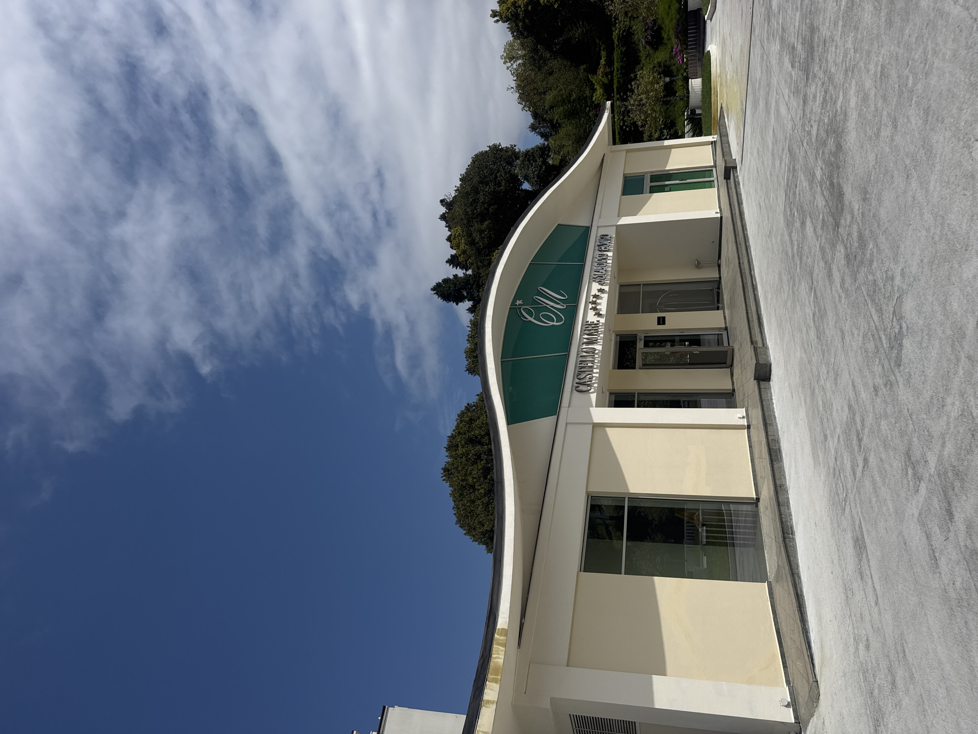 The image captures the exterior of a modern, two-story building under a clear blue sky with sparse white clouds. The building features a distinctive, curved roofline and is painted in a pale yellow color with prominent green accents on its upper facade, where the main entrance is located. A stylized "CM" logo with a star is visible on the green section, along with the text "CASTELLO MARE" and below it, "*** ALL-SUITES RESORT." The building has multiple windows with dark frames, reflecting the surroundings. In front of the building is a wide, light gray paved area, resembling a driveway or pedestrian path. Lush green trees and foliage are visible behind and to the sides of the building, suggesting a well-landscaped or natural setting. The scene is static, showing no activity or people. The bright lighting indicates it is daytime, likely morning or early afternoon, with distinct shadows cast on the paved surface.