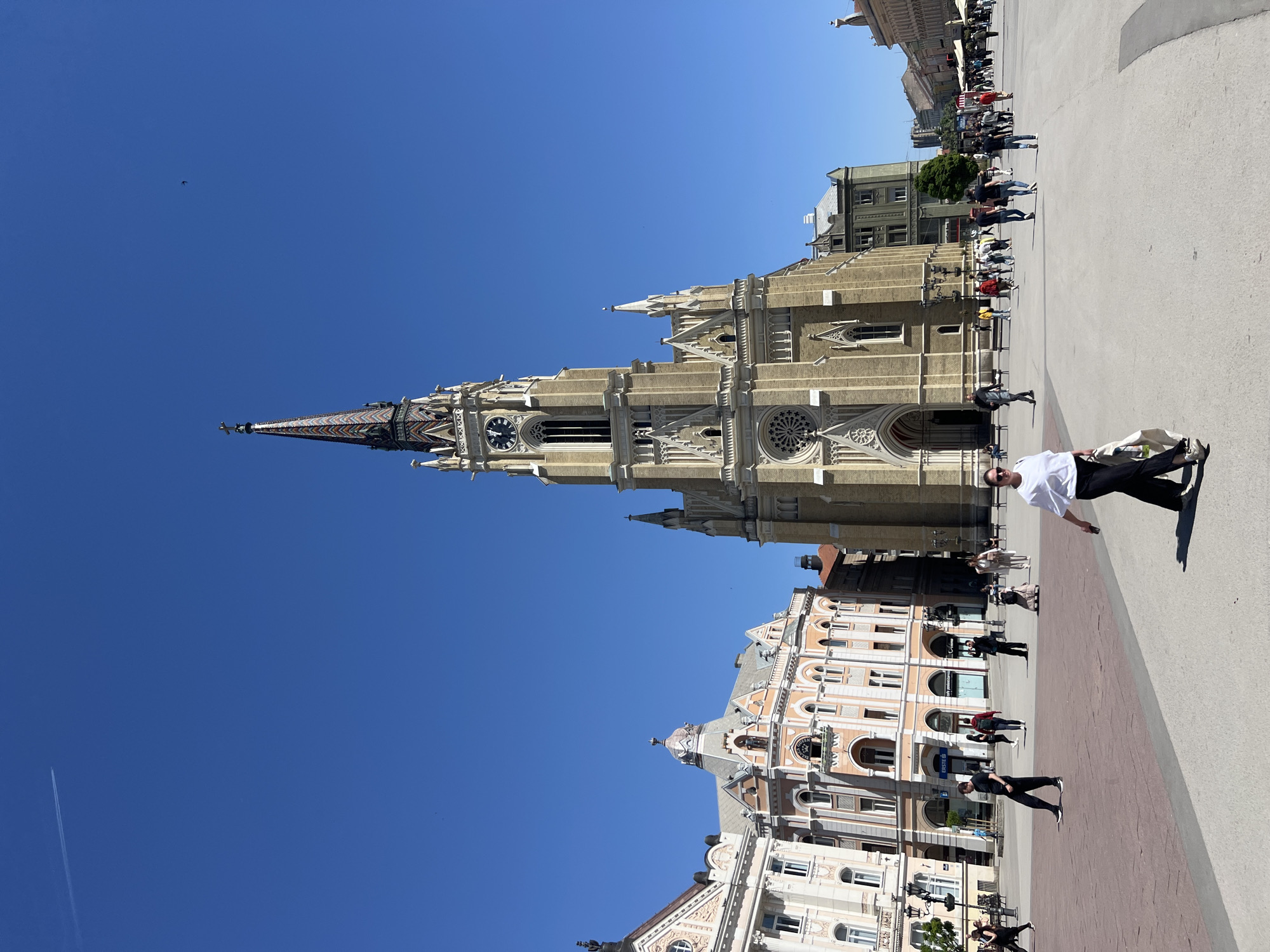 The image shows a view of the Freedom Square in Novi Sad, Serbia. The Roman Catholic Church of the Name of Mary is the prominent building. Several other buildings are visible, alongside a plaza with people walking around. One person wearing a white shirt, dark pants, and sunglasses stands out in the foreground. The weather is sunny, with a clear blue sky.
