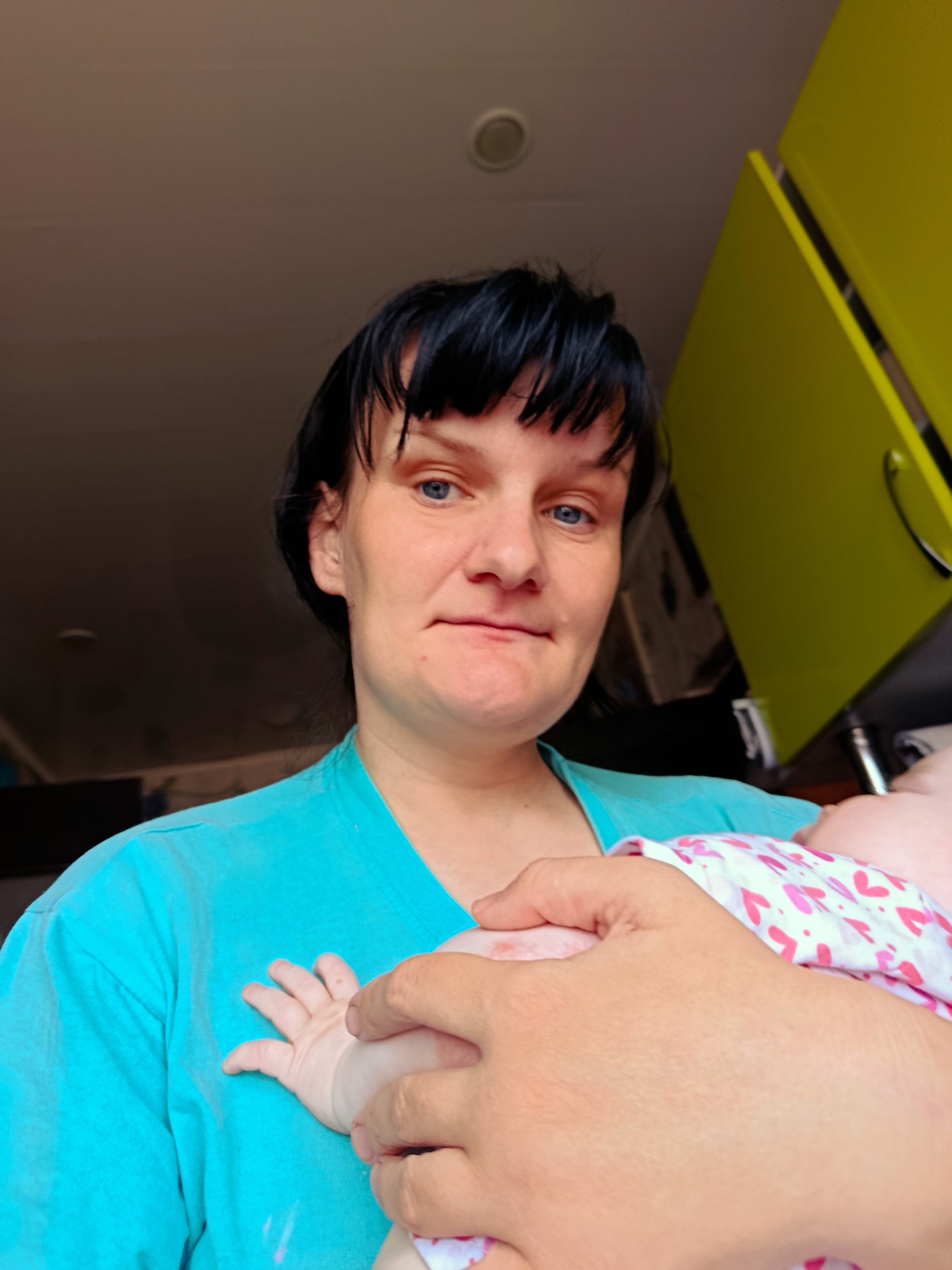 A woman with short, dark hair and blue eyes holds a baby wrapped in a white blanket with pink hearts. She wears a turquoise t-shirt. The background includes a ceiling with a round light fixture and yellow kitchen cabinets. The location cannot be determined from the image, and the city of Фоки, Russia, is not discernible.