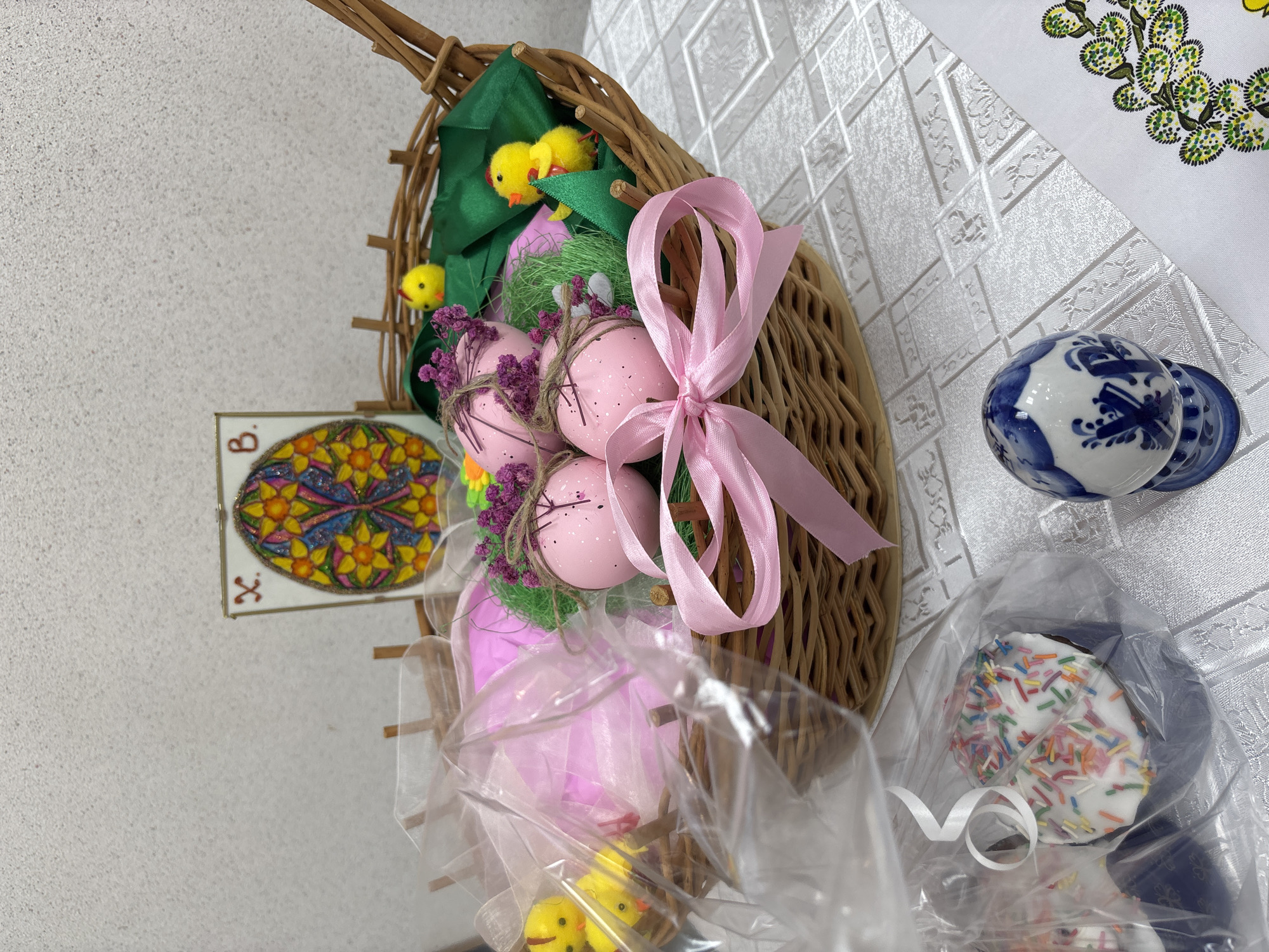 A tilted eye-level shot depicts an Easter basket on a white patterned surface. The basket, made of woven material, contains pink eggs decorated with twine and purple flowers, green foliage, and small yellow chicks. A pink ribbon is tied to the basket. Beside the basket, resting on the surface, is a blue and white patterned egg and a wrapped dessert with white frosting and colorful sprinkles. To the left, on a light-colored wall, hangs a framed image with a stained-glass-like design and a letter "B" in the upper left corner. The image may be located in Perm, Russia.