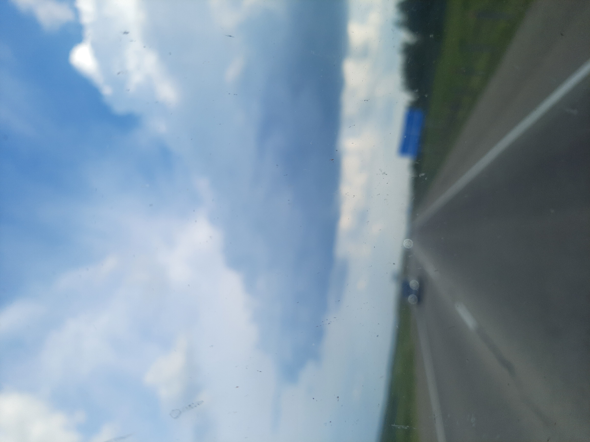 The media file captures a perspective from inside a vehicle, looking outwards through the windshield. The scene depicts a highway stretching into the distance, with lush green fields and trees lining the right side. The sky is a prominent feature, filled with dramatic, swirling white clouds against a vibrant blue backdrop, suggesting it is daytime with fair weather. A blue road sign is visible on the left side of the road. The image is slightly out of focus and has some dirt or smudges on the glass, giving it a slightly hazy appearance. The road itself has white lane markings. The overall impression is that of a journey on a clear day, with the focus being on the expansive sky and the open road ahead.