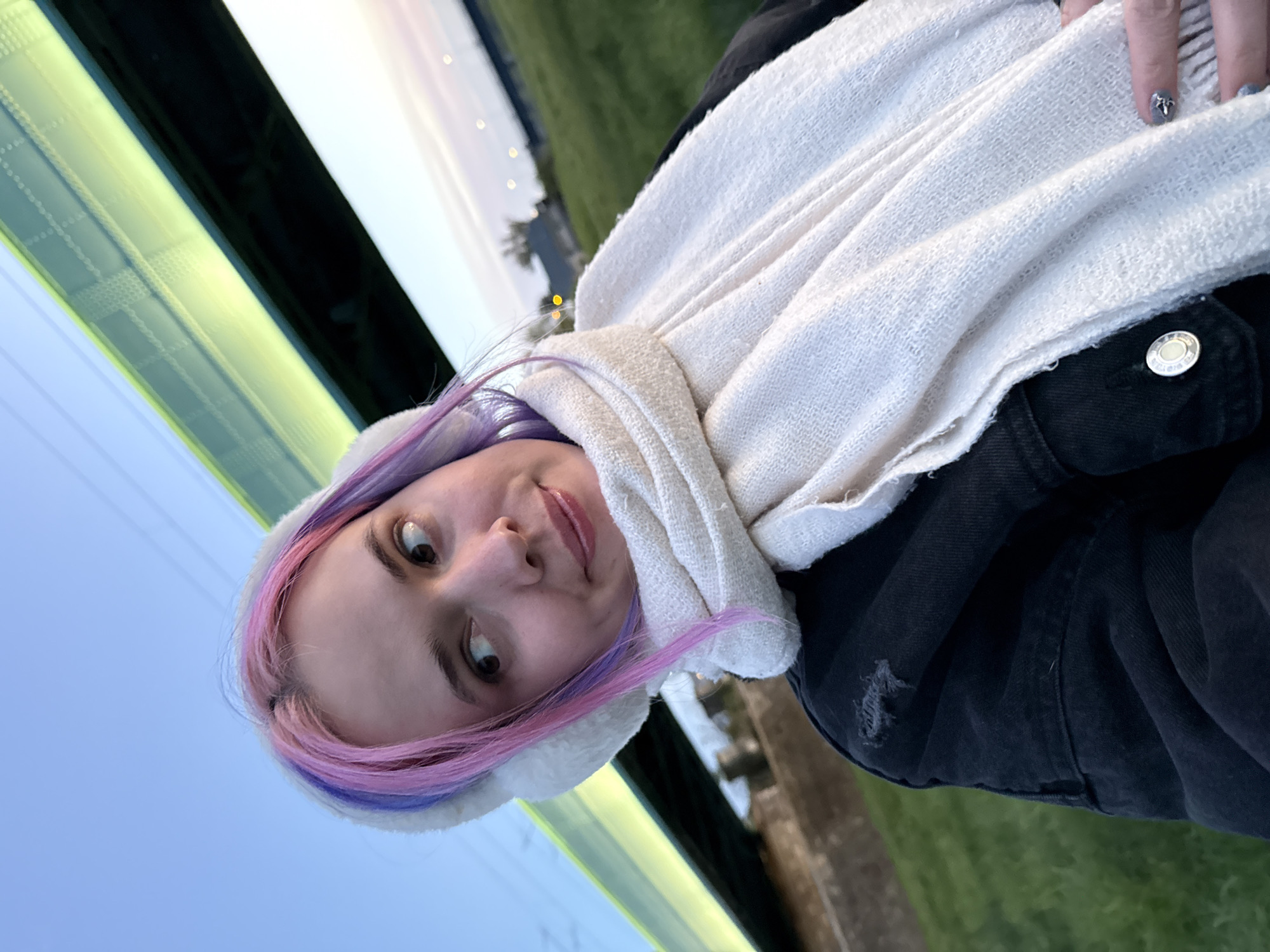 A woman with dyed purple and pink hair is wearing a white ear-covering hat and a white scarf. She is also wearing a black jacket. The background shows a blue sky and green grass. A structure with green lights is visible. Based on the environment, it is likely a public transportation shelter or building. The location is presumed to be the city of Zagreb, Croatia.