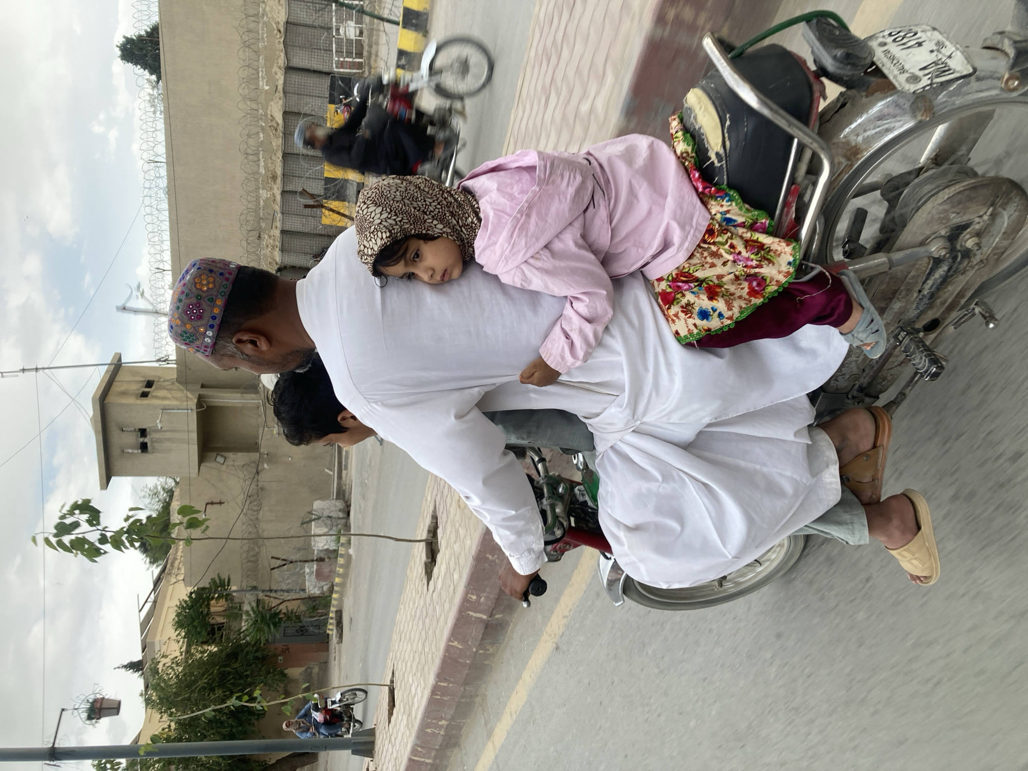 The image, taken in Quetta City Tehsil, Pakistan, depicts a daytime urban scene. A man is riding a motorcycle, seen from behind, wearing a white shalwar kameez and a distinctive, embroidered colorful cap. A small child, likely a girl, rests or sleeps comfortably against his back, wearing a pink jacket, a patterned headscarf, and maroon pants, with a floral print visible on her clothing. Another person is partially visible between the man and the child on the motorcycle.
The silver-colored motorcycle has a black seat, and its license plate clearly displays "NHA 488 PUNJAB 188". The scene unfolds on a paved road with a brick sidewalk to the right. In the background, light-colored buildings with barbed wire along their walls are visible, under an overcast sky. Other vehicles and pedestrians can be seen in the distance, indicating a bustling street. The image captures a quiet moment of everyday travel, with the child peacefully resting amidst the urban activity.