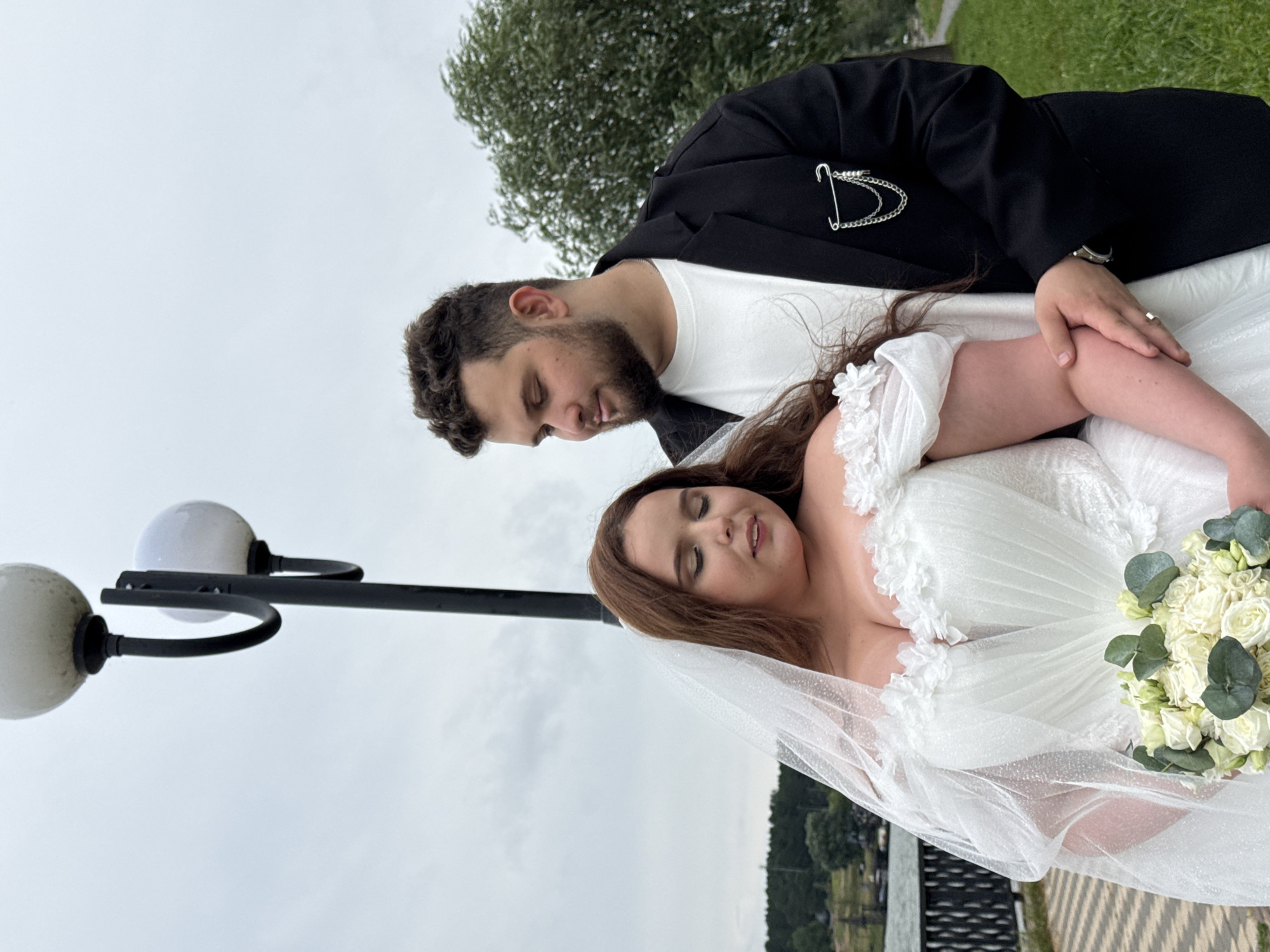 In Votkinsk, Russia, a wedding photo captures a couple standing outdoors with a cloudy sky overhead. The bride, with long, auburn hair and a white wedding dress featuring ruffled off-the-shoulder sleeves and a sweetheart neckline, is holding a bouquet of white roses and eucalyptus. Her veil, edged with glitter, drapes over her. The groom, wearing a black suit jacket over a white t-shirt and a decorative chain with safety pins on the lapel, stands beside her, his arm around her waist. They are positioned near a lamppost with two globes. In the background, a body of water, a decorative fence, a path, and lush greenery are visible.