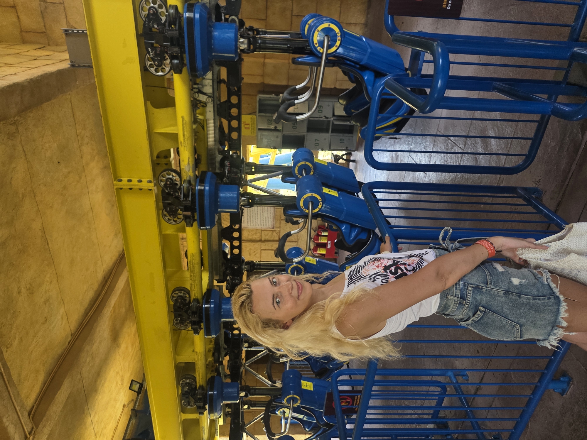 A blonde woman in a white tank top and denim skirt is seated in the blue metal seat of a ride at an amusement park. The ride features a yellow beam with blue mechanisms and wheels. The environment appears to be indoors with stone-like walls in the background. The location is Đà Nẵng, Vietnam.