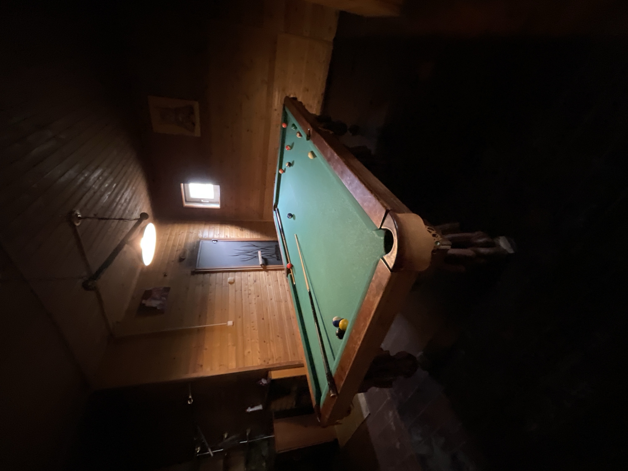 The image depicts an indoor room with a pool table dominating the scene. The pool table is green and brown with several billiard balls on the surface. Two cues are also present. The room has wooden walls and a low ceiling. A window and a picture frame are visible on the wall. The exact location in Kommunarka, Russia, is unknown as no specific identifiers are visible.