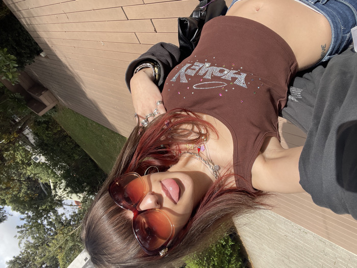 An upward-angled selfie depicts a female individual outdoors in Raanana, Israel. The subject, viewed from the chest down to the upper thighs, has long brown hair with reddish highlights and bangs, and wears large, dark reddish-brown sunglasses. Her tongue is extended from her mouth. She is dressed in a dark brown sleeveless crop top with "HONEY" spelled out in silver-colored embellishments, encircled by a similar oval design. Multiple layered silver-colored necklaces with various charms are visible around her neck. Her right hand, visible on the left side of the image, displays several silver-colored rings, and her right wrist wears multiple bracelets. A dark grey or black garment is draped over her shoulders. Dark blue jeans are visible at the lower left, exposing her midriff and a tattoo reading "LOVE" on her left hip. The foreground consists of a light brown wooden-slat or composite decking surface. Beyond this, a manicured green lawn is visible, backed by dense green foliage from trees and bushes. Portions of light-colored buildings are discernible through the foliage in the upper background. The scene is illuminated by direct sunlight, casting shadows.