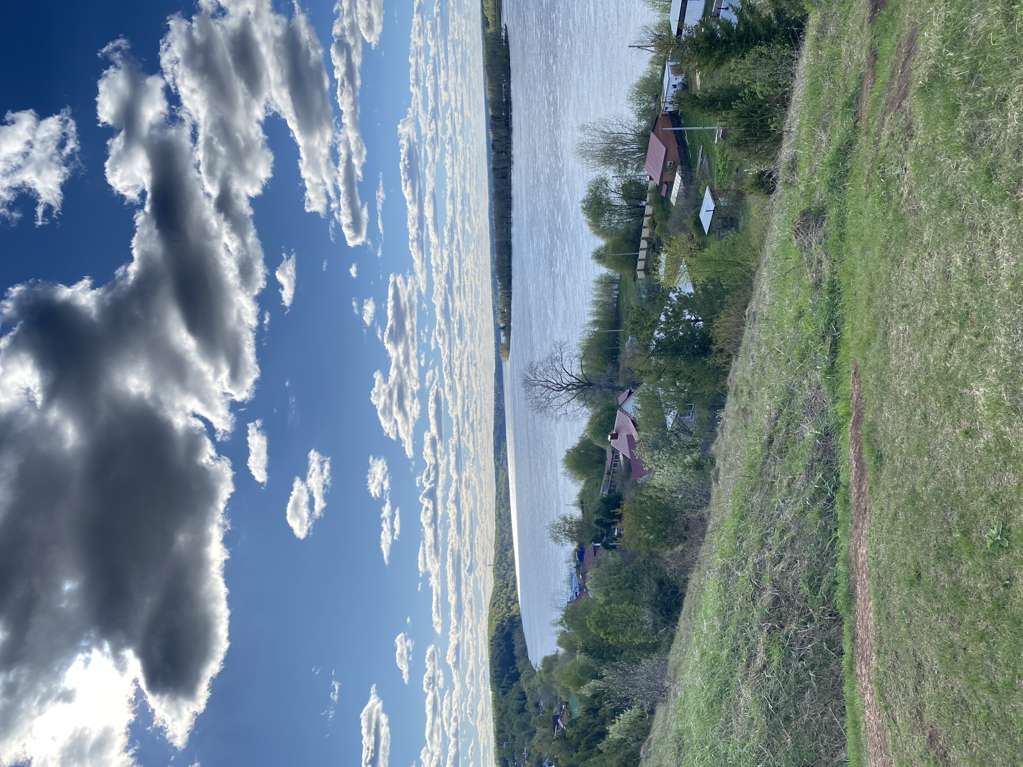 From an elevated vantage point in Ольховка, Russia, the landscape is captured. A wide river dominates the center of the frame, reflecting sunlight. On one bank, a dense forest stretches along the river's edge. The opposite bank features a mix of trees and residential structures, including houses with red roofs. Grassy terrain occupies the foreground. A blue sky dotted with cumulus clouds is visible above.