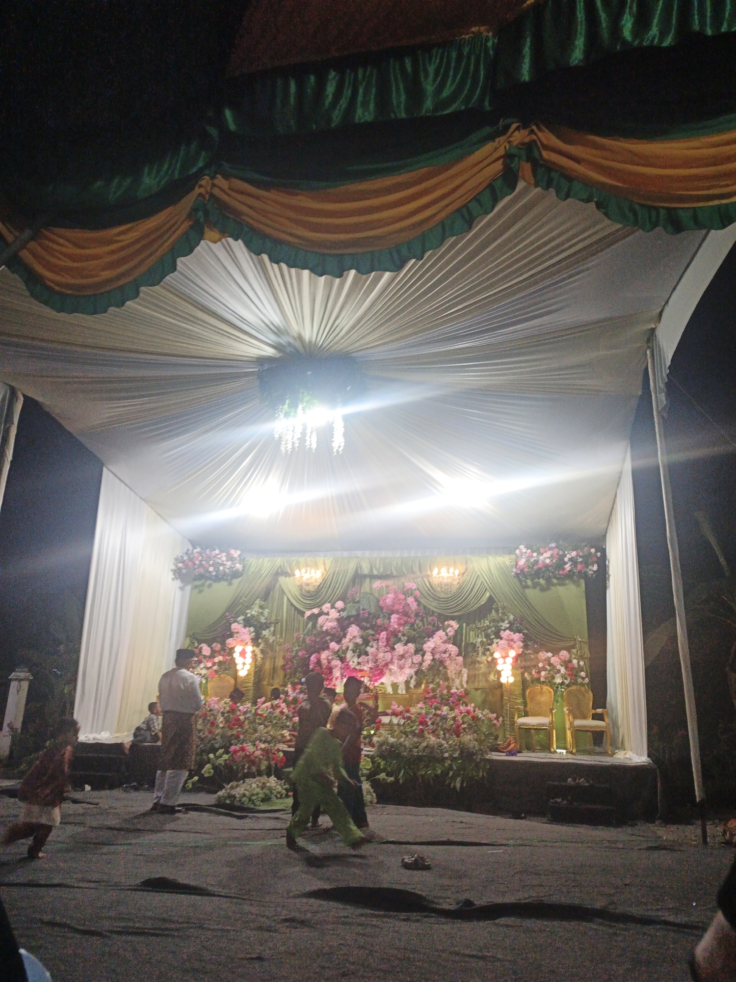This is a medium shot, taken at night, of an outdoor event space decorated for a celebration. The scene appears to be located in Pematang Duku, Indonesia, as suggested by the context. The primary focus is a tented area with elaborate drapes of white, gold, and green fabric. A chandelier hangs from the white ceiling.
The background features a stage area adorned with an abundance of pink and red flowers, draped green fabric, and two ornate gold chairs. Lighting fixtures, possibly candelabra, are present on the stage. White curtains line the sides of the tent.
In the foreground, several people are visible. To the left, a young child in a reddish-brown outfit is running towards the right. In the center, a person in a white shirt and patterned sarong stands facing away from the camera. To their right, two children, one in a green outfit and another in darker clothing, are in motion, possibly playing. Another adult is partially visible in the background to the right, standing near the stage. The ground is covered in a dark, textured material, likely an outdoor carpet or tarp. The overall atmosphere suggests a festive gathering.