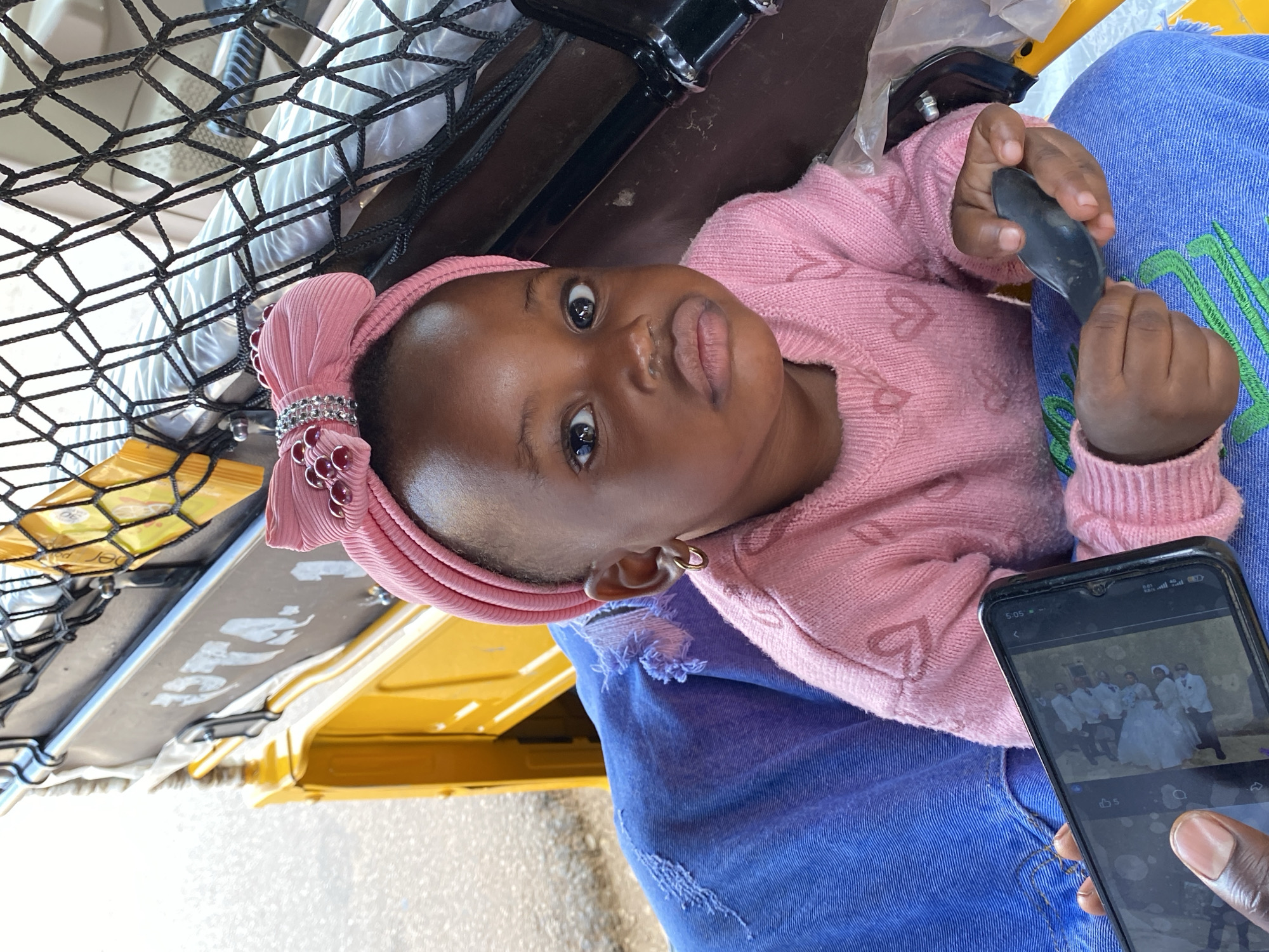 The image depicts a baby girl lying in what appears to be a vehicle, possibly an auto-rickshaw, judging by the yellow paint and the metal framework visible. She wears a pink sweater with heart patterns and a pink headband with a bow. She is holding a small, dark object in her hands. The background suggests a bright, outdoor setting. A mobile phone displaying a photo of a wedding is in the bottom right corner. The location is suspected to be Vom, Nigeria.