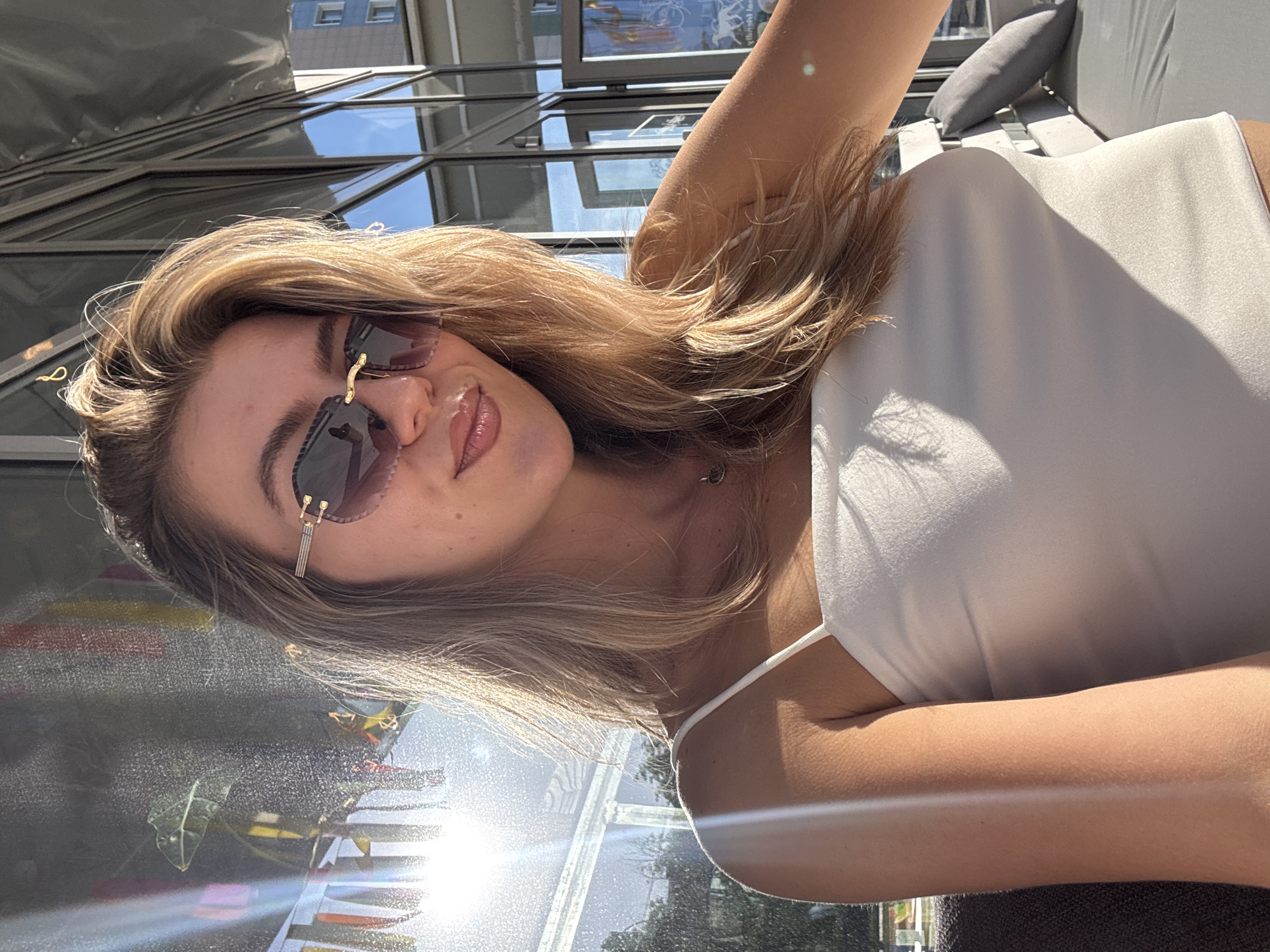A young woman with blonde hair and sunglasses is pictured in a selfie, wearing a white tank top. She is in an outdoor urban setting, possibly a patio or balcony, with glass windows or doors reflecting the city. Part of a grey cushion and outdoor furniture are visible in the background. The location is in an unknown city.