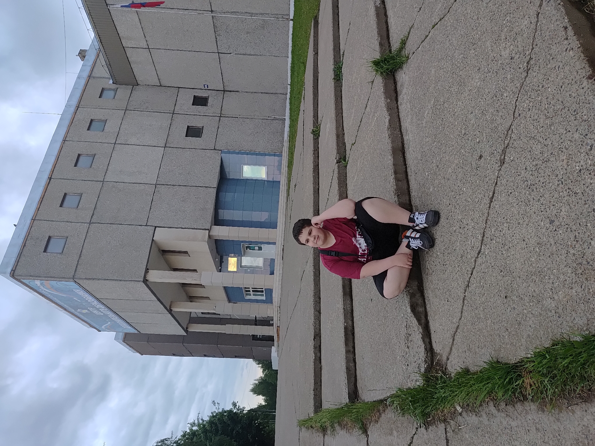 A young person sits cross-legged on a concrete slab sidewalk in Марковский, Russia. They wear a maroon t-shirt, black shorts, and black sneakers with white stripes. The person is looking off-camera with a hand near their head. Behind the person is a large, multi-story grey building with square windows, a blue-tiled entrance area, and a sign with Russian text on the left side. The sky is overcast, and trees are visible in the lower left corner. Grass grows in the cracks of the concrete sidewalk.