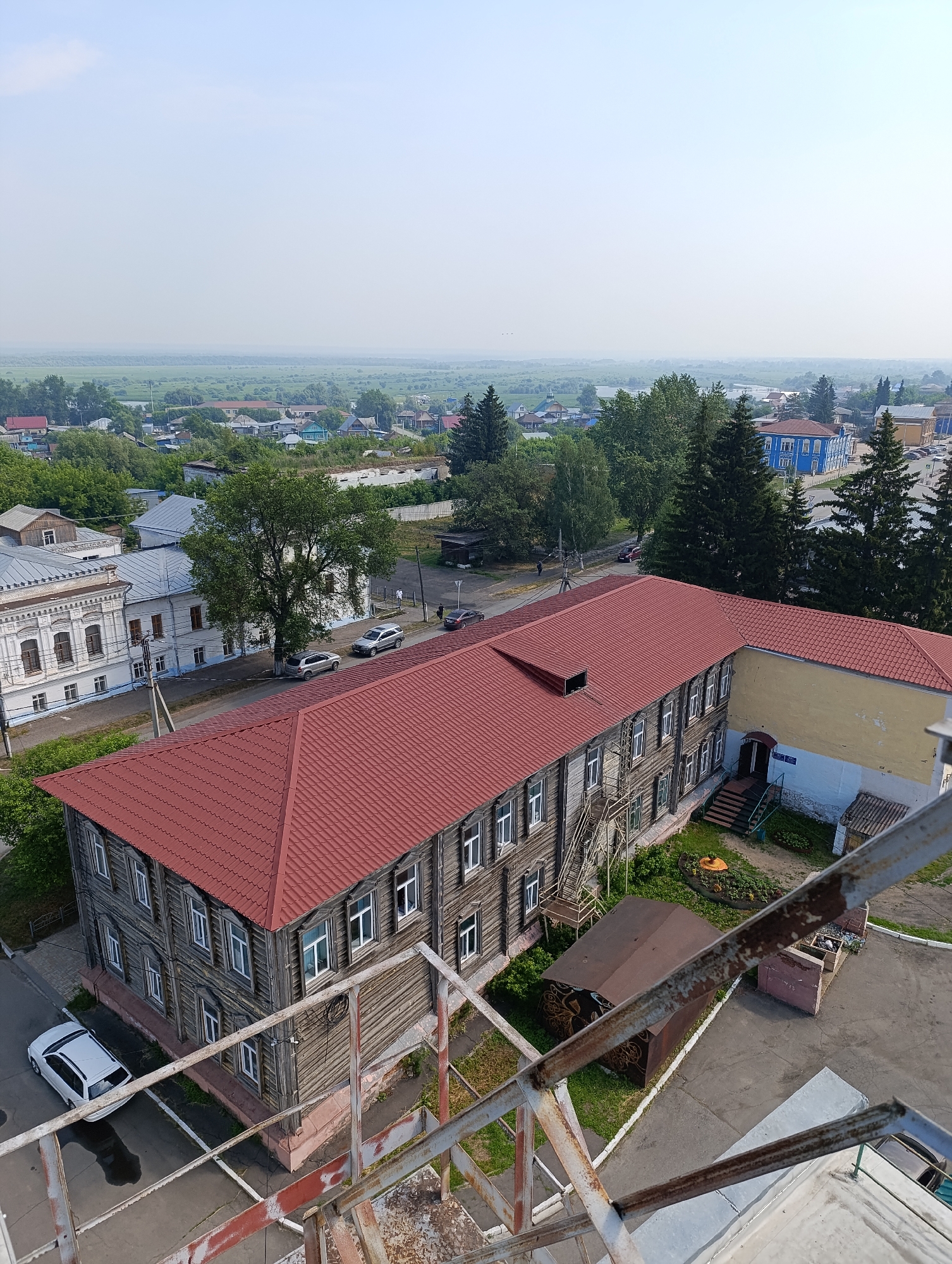 This is an aerial, wide-angle shot of the city of Tara, Russia, on a clear, bright day. The image captures a landscape of predominantly low-rise buildings, interspersed with trees and green spaces. In the foreground, a large, two-story wooden building with a red-tiled roof dominates the scene. This building features numerous windows and an external staircase on one side. Adjacent to it is a modern, yellow and white building. A white car is parked on the street in front of the wooden building. Further in the distance, more houses and buildings of varying architectural styles are visible, along with a vast, flat, green landscape stretching towards the horizon. The sky is a pale blue, with a slight haze. The image appears to be taken from a high vantage point, possibly a tower or a taller building.