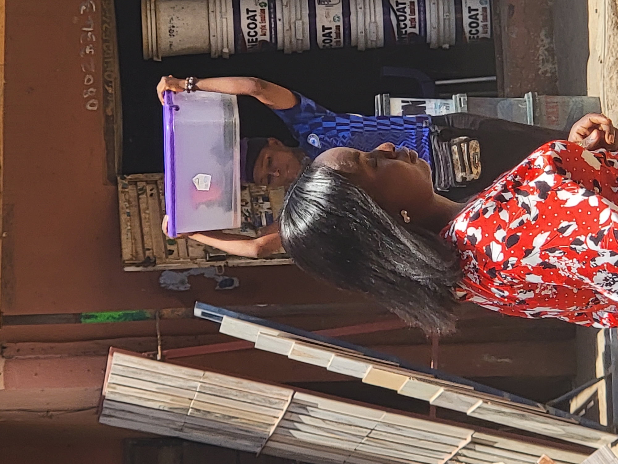 In Maiburiji, Nigeria, a woman in a red and white patterned blouse and a man in a blue Chelsea FC shirt are visible. The man holds a translucent plastic container with a purple handle. A wall with painted white numbers and a line of white paint buckets labeled "ECOAT Acrylic Emulsion" are in the background. Part of a slatted blind or awning is in the foreground.