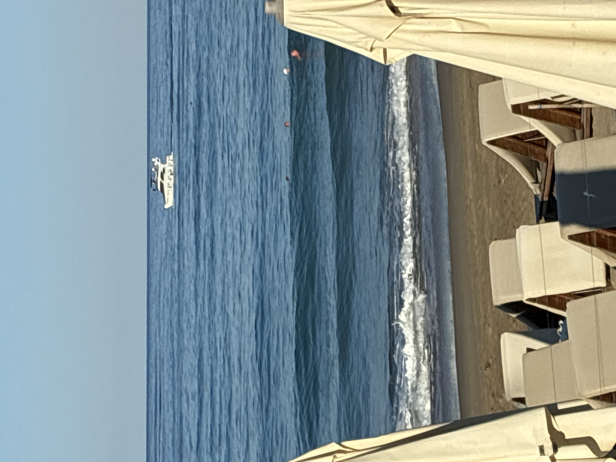 The image captures a view of the Mediterranean Sea from a beach in Alanya, Turkey. In the distance, a white yacht navigates the calm blue water under a clear sky. Closer to the shore, several individuals are visible swimming or wading in the sea, their heads appearing as small dots. The foreground of the image is dominated by beach chairs and large, cream-colored umbrellas, suggesting a resort or public beach setting. The sand is visible between the chairs, and gentle waves break along the shoreline. The overall scene conveys a peaceful and sunny day at the beach.