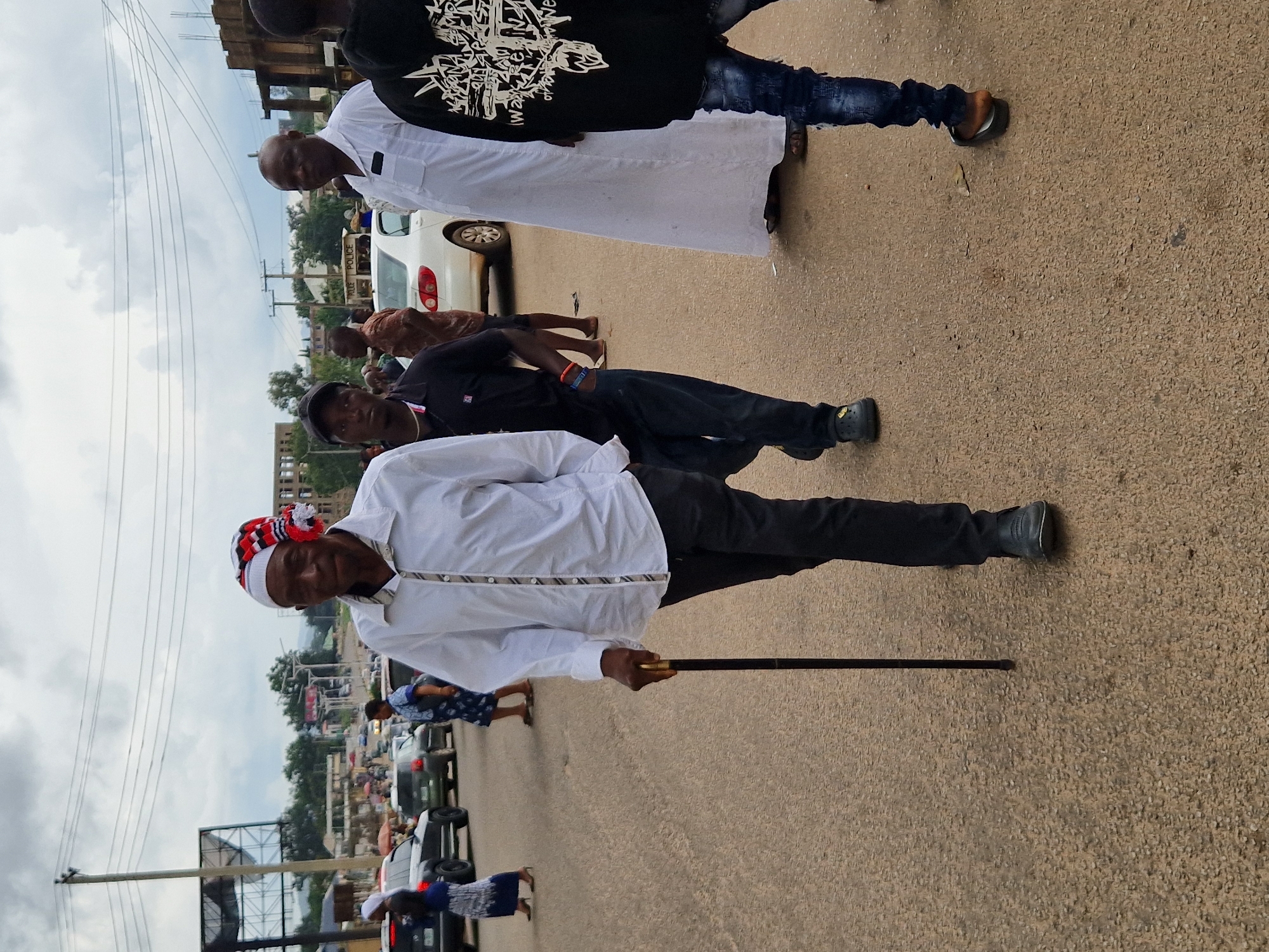 The image depicts a street scene in Kabong, Nigeria, with several individuals walking on a paved road. In the foreground, an older man with a distinctive red, white, and black patterned head covering, walks with a cane. He is wearing a white long-sleeved shirt and dark trousers. To his right, a man in a black polo shirt and dark trousers walks beside him. Further in the background, other individuals are visible, some walking, others standing. Cars and a white building with scaffolding are present in the background, suggesting an urban environment. The sky is overcast with clouds.