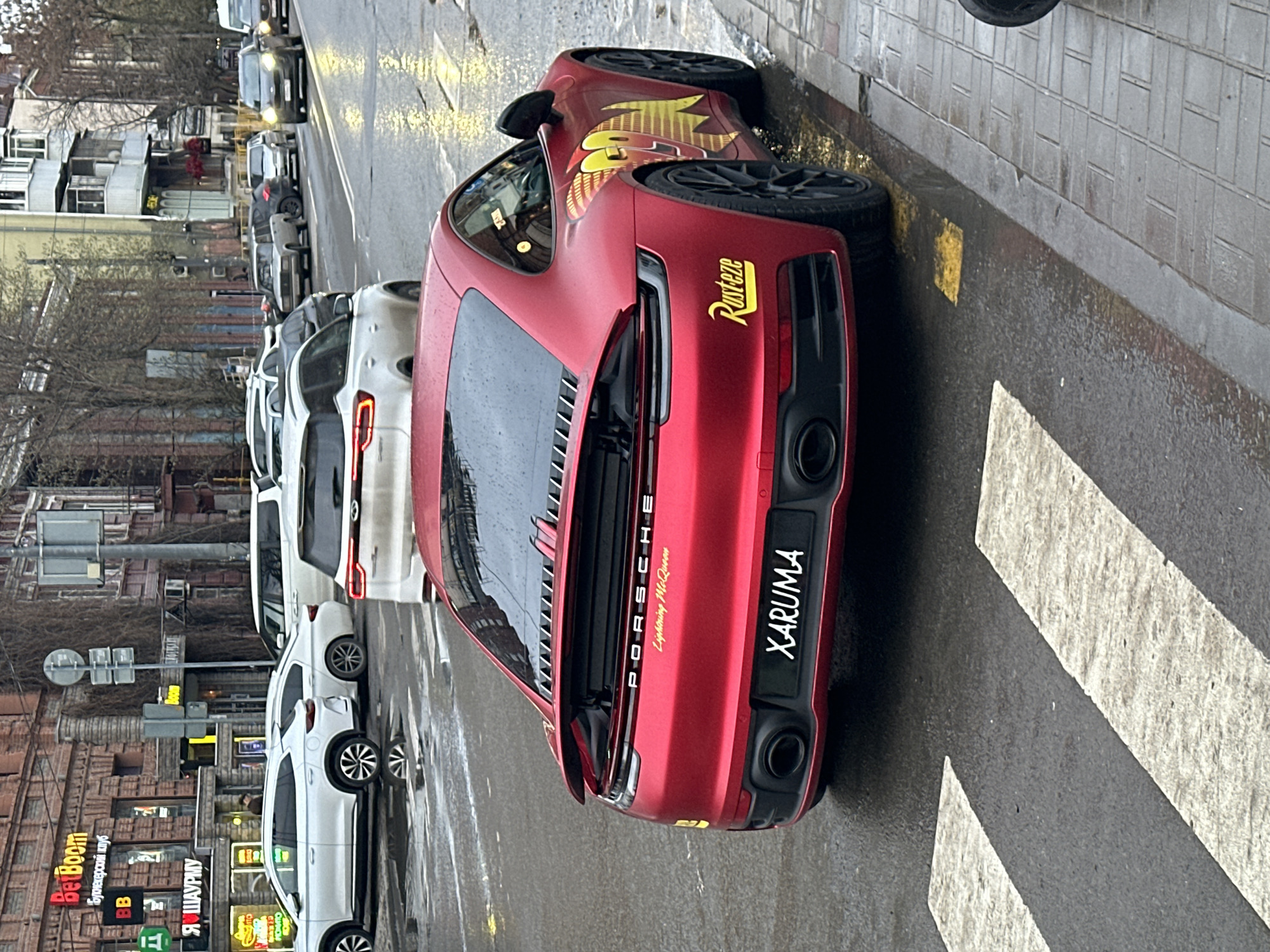 A red Porsche with "Rusteze" and "Lightning McQueen" markings is parked on a street in Rostov-on-Don, Russia. The license plate reads "XARUMA". The car is parked near a pedestrian crossing, with white cars parked behind it. A building with signs for "BetBoom" and "Я ШАУРМУ" is visible in the background. The street appears to be wet.