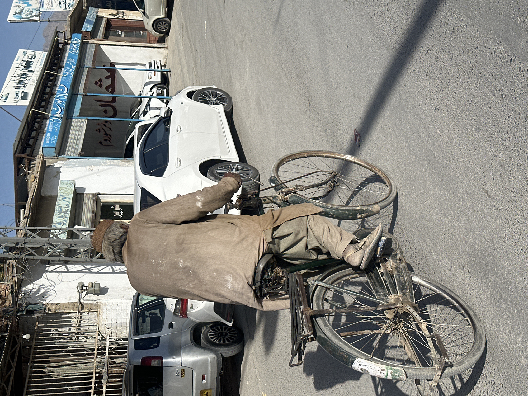 The image displays a street scene with an overhead perspective, looking down at a man riding a bicycle. The man, possibly elderly, has gray hair under a light brown cap and wears a dusty, light brown traditional garment. He is pedaling an old, dark green bicycle with visible rust on the spokes and what appear to be worn or deflated tires.
The setting is an urban or semi-urban environment. On the right side of the frame, a modern white car is parked on the street, and behind it, a silver older model van or SUV is partially visible, displaying "K-662" on its rear bumper sticker. Buildings line the left side of the street, featuring various storefronts. One building has a prominent blue awning with text in an Arabic script, including characters that resemble "تشغيل" (Tashghil - operation/start) or a similar word. Further text in the same script is present on other signs and awnings, though difficult to decipher fully. Barbed wire is visible on a wall near some metal scaffolding. Mountains can be seen in the distant background, rising above the buildings.
The lighting suggests bright daylight, casting strong shadows on the asphalt road surface. The overall impression is of a practical, bustling street scene, likely in a South Asian or Middle Eastern country, given the script, architecture, and vehicles.