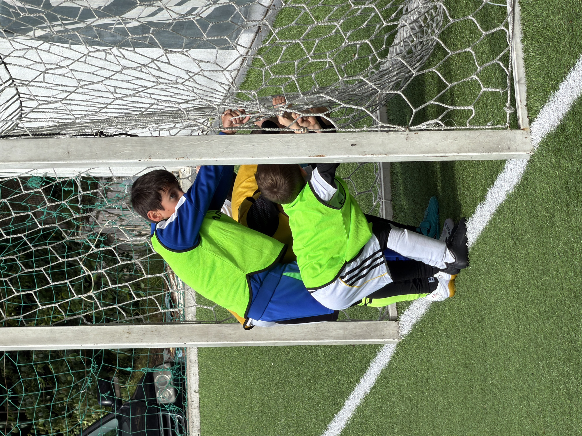 Here's a concise description of the image:
The image depicts several children entangled within a soccer goal on a grassy field. The location appears to be Sochi, Russia. Several of the children are wearing brightly colored soccer practice vests (one green, one yellow), while others are in soccer uniforms. The children are positioned between the goalposts and netting, with some holding onto the net. The setting appears to be outdoors on a soccer field or practice area, with green grass and white lines visible. The scene suggests a playful or perhaps accidental entanglement of the children within the soccer goal. In the background, partial view of trees, a vehicle (possibly a car) and an overhead electric wire can be seen.