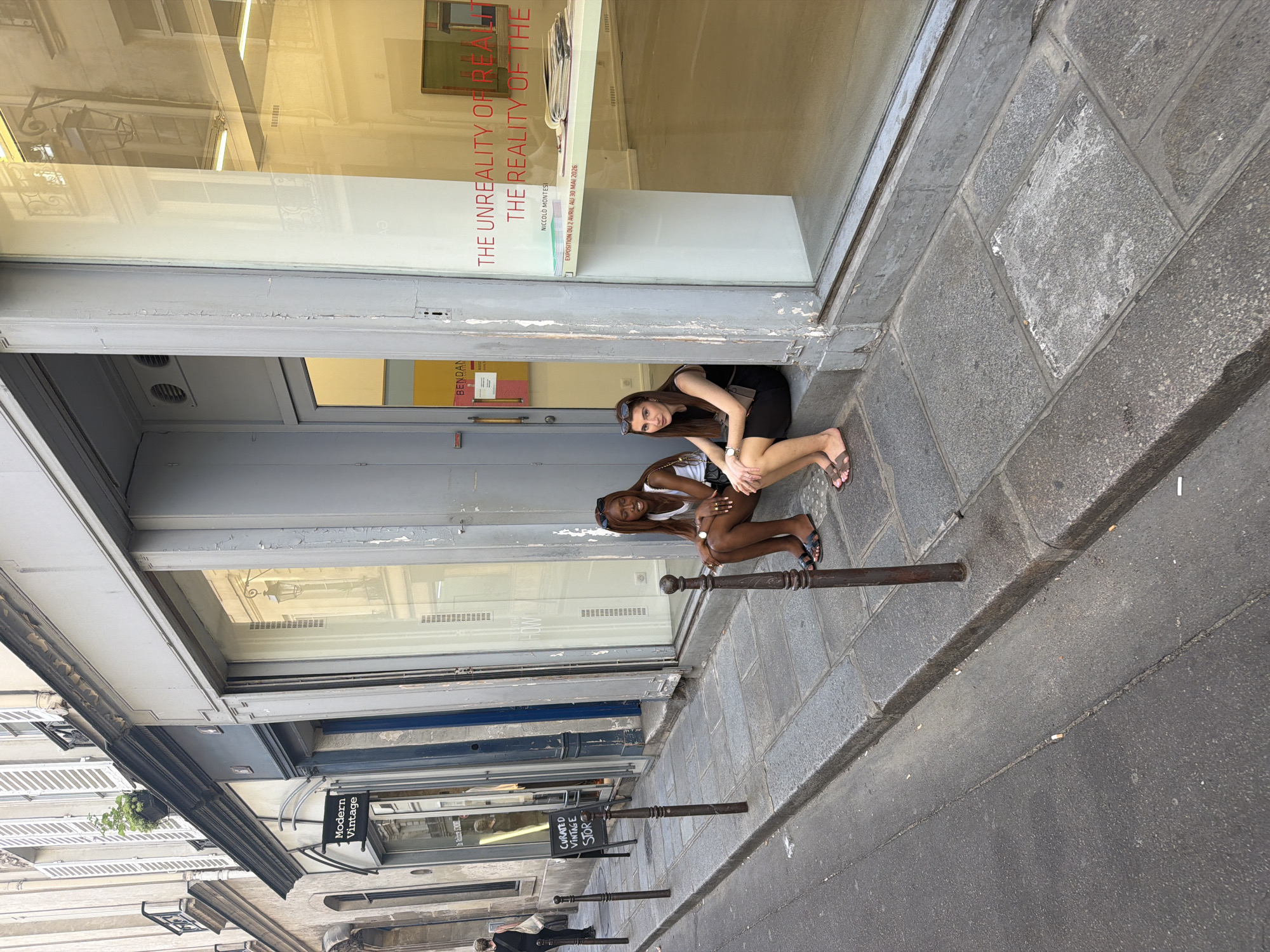 The photograph shows two young women crouching down in front of a storefront in Paris, France. The women appear to be posing for the photograph. The storefront is lined with windows, and a sign reads "THE UNREALITY OF REA THE REALITY OF THE", with "NICCOLO MONTESI" and an exposition date below it. A sign for "Modern Vintage" is visible on a nearby storefront. Metal bollards line the street in front of the storefronts. The street appears to be constructed with gray stone pavers.