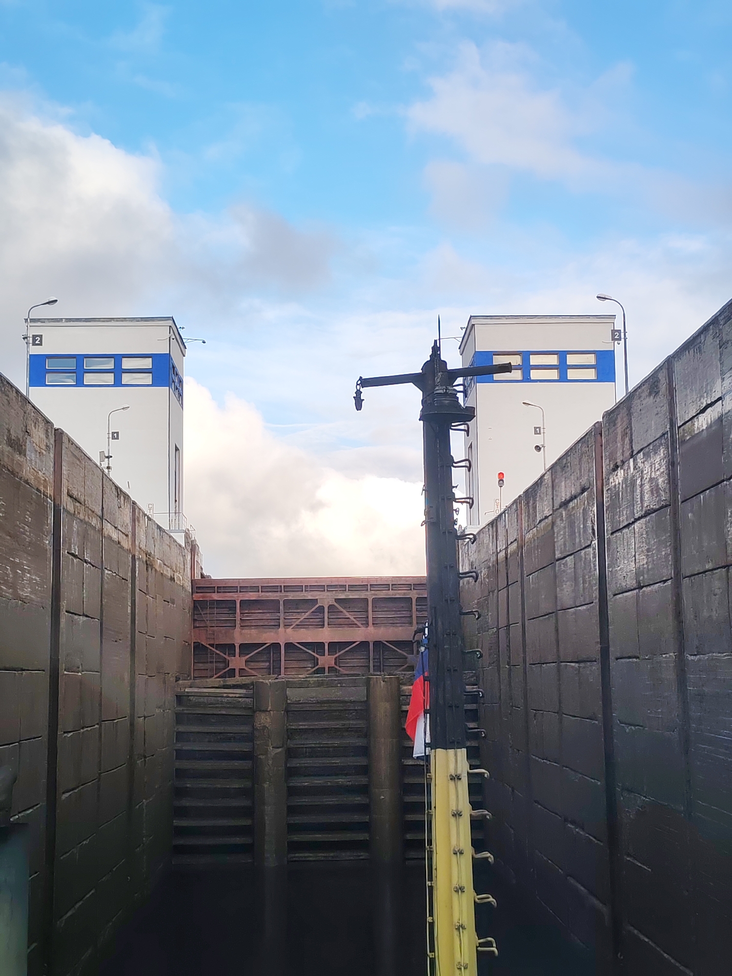 The image captures a view from inside a lock on a waterway, potentially a canal or river. Concrete walls define the lock's sides, appearing worn and marked with water lines. At the end of the lock, a gate structure is visible. On either side of the gate stand tower-like structures with light-colored exteriors and blue accents around windows. A utility pole with a yellow ladder and a flag attached is present in the foreground. The sky is visible above the towers, showing a mix of blue sky and clouds. The location is stated as "Unknown, Unknown". The water level within the lock is relatively low, exposing stepped sections along the lock's walls.