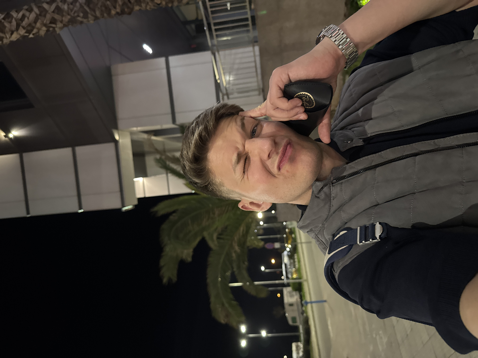 A man is taking a selfie at night. He is wearing a gray vest and a watch. He is holding a Ray-Ban case to his face and winking. The location appears to be an outdoor area at night in the city of Plovdiv, Bulgaria. The background features palm trees, streetlights, and a modern building.