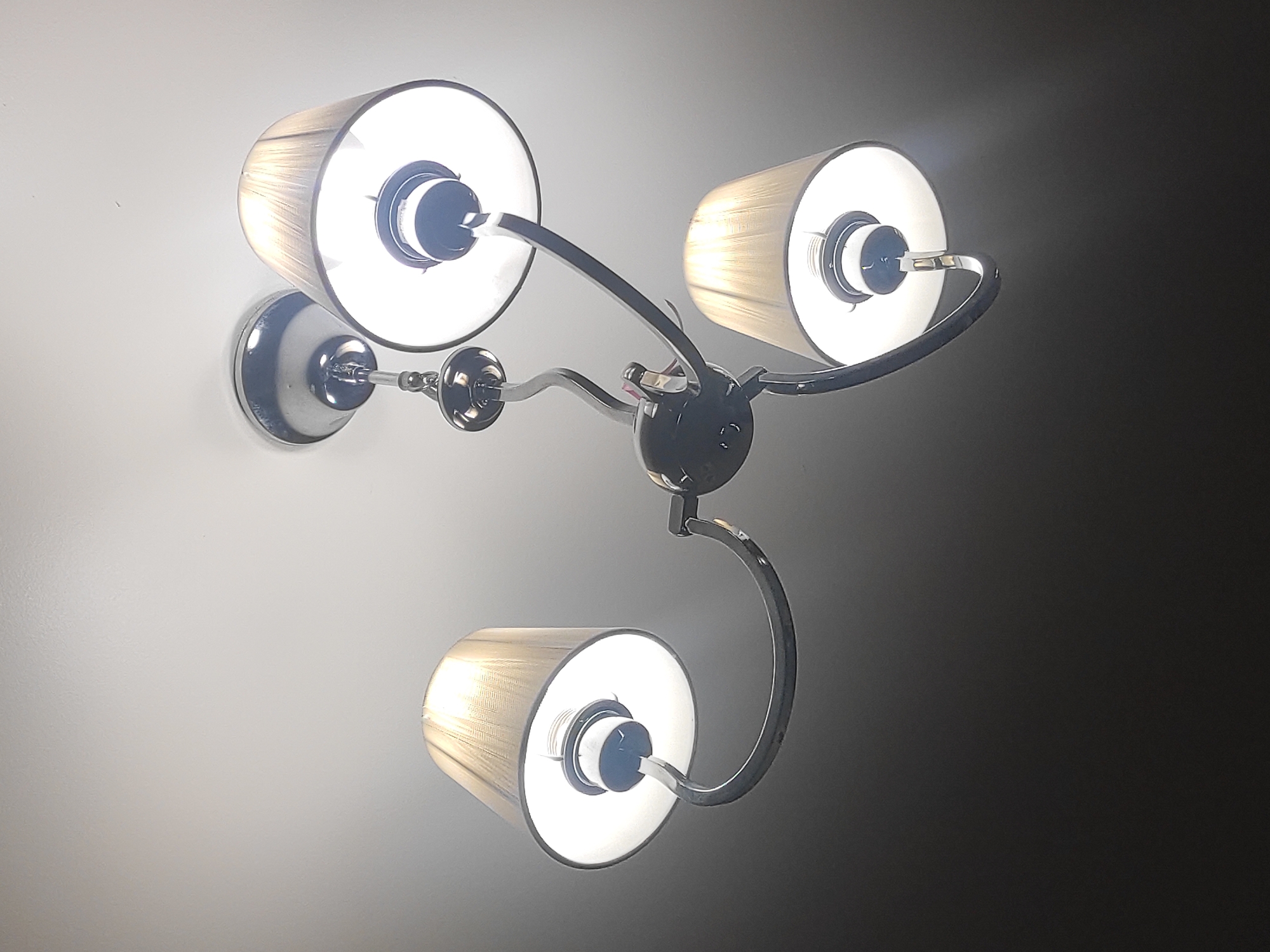A modern, three-armed light fixture is prominently displayed, likely mounted on a wall or ceiling indoors. The fixture features a polished chrome or silver-finished central base from which three slender, curved metal arms extend. Each arm supports a cylindrical lampshade, which appears to be made of a textured, possibly fabric, material with subtle pleats or ribs, in a light color such as white or cream.
Within each lampshade, a bright light bulb is illuminated, emitting a cool-white light that brightly illuminates the immediate area around the fixture. The light creates a strong contrast against a darker background on the right side of the image, while the area to the left of the fixture is brightly lit, suggesting a light-colored wall or ceiling. The scene depicts the light fixture in active use, providing illumination. No people, discernible activities beyond the fixture being on, or visible text are present in the image. The ambient lighting suggests an interior setting where artificial light is necessary, possibly indicating evening or nighttime.