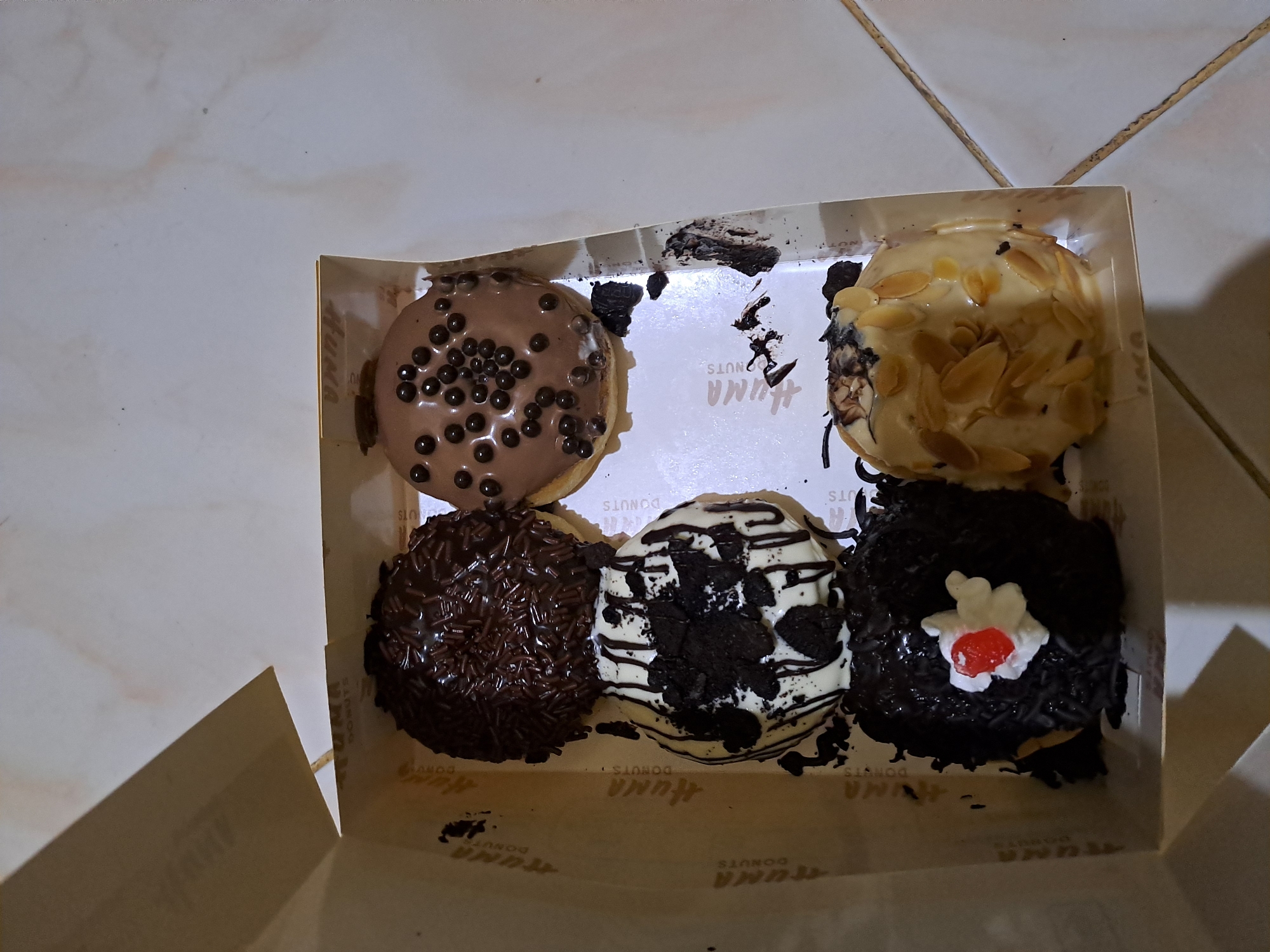 Six donuts are arranged in a white cardboard box, which is open and placed on a light-colored tiled floor. The donuts are positioned in two rows of three.
In the top row, the donut on the left is covered in dark chocolate sprinkles. The donut in the center of the top row has a light brown glaze and is topped with small, dark brown spherical candies. The donut on the right of the top row is partially visible and appears to have a light brown glaze.
In the bottom row, the donut on the left is dark brown with white frosting drizzled on top, and pieces of dark cookie crumbs. The donut in the center of the bottom row is dark brown and decorated with dark sprinkles and a small white frosting topping with a red cherry in the middle. The donut on the right of the bottom row is covered in a pale yellow glaze and topped with sliced almonds.
The box has the "HUNA DONUTS" logo printed on the inside. The surrounding environment is a tiled floor. No people are visible in the image. The lighting suggests an indoor setting, likely a home or a food establishment. The overall scene is a simple, straightforward presentation of donuts in a box.