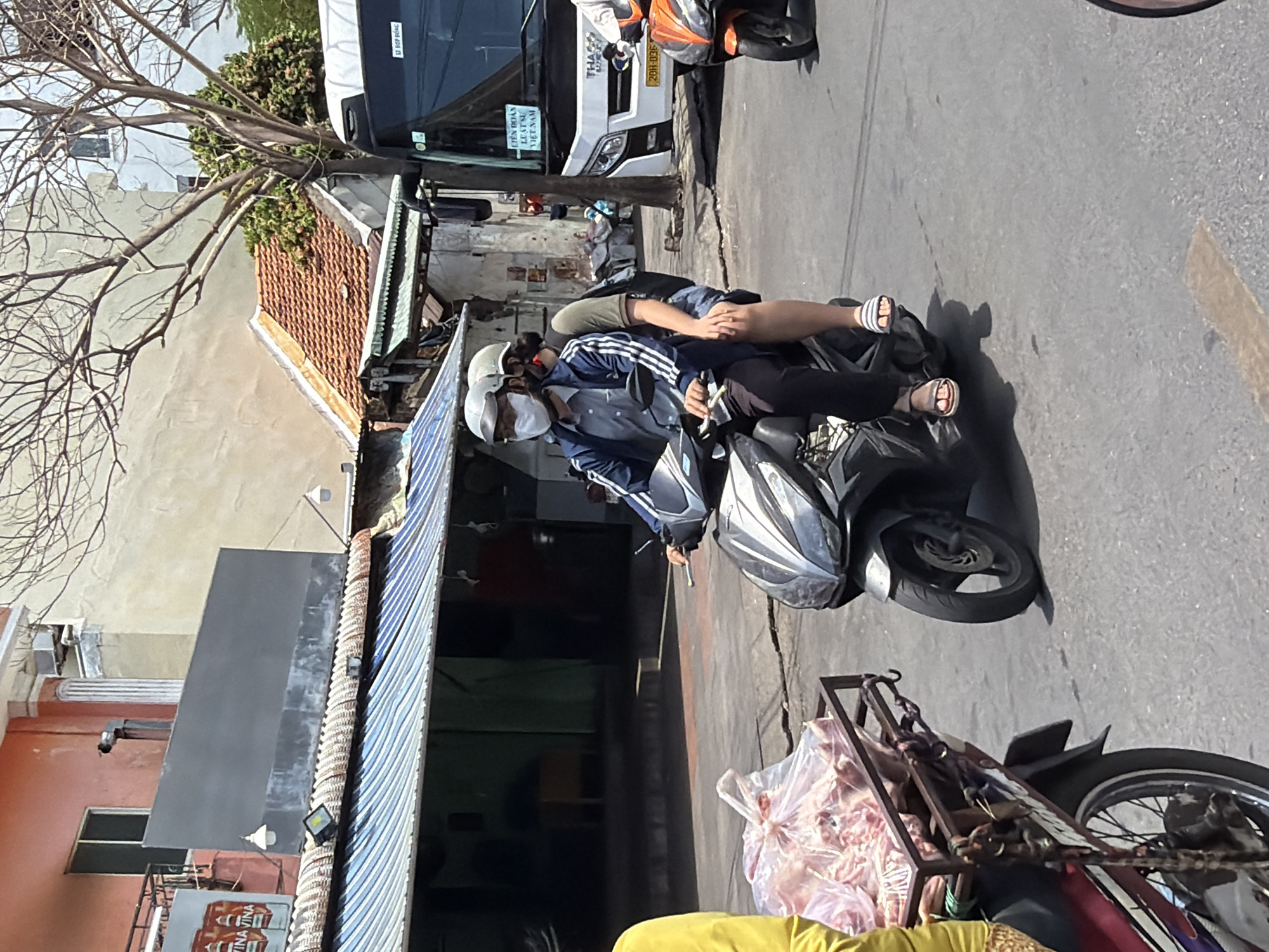 The image depicts a street scene in Nha Trang, Vietnam, on a sunny day. Two individuals are riding on a scooter, a man in the front wearing a face mask and a blue jacket with white stripes, and a woman behind him. The man is holding the handlebars, and the woman has her legs resting on the scooter. The scooter is silver and black. In the background, there is a white bus with signage that appears to be in Vietnamese, and another orange scooter is visible to the right. To the left, a person in a yellow shirt is partially visible, next to a metal rack loaded with plastic bags containing meat or animal products. Buildings line the street, with awnings and tiled roofs. The road surface is asphalt.