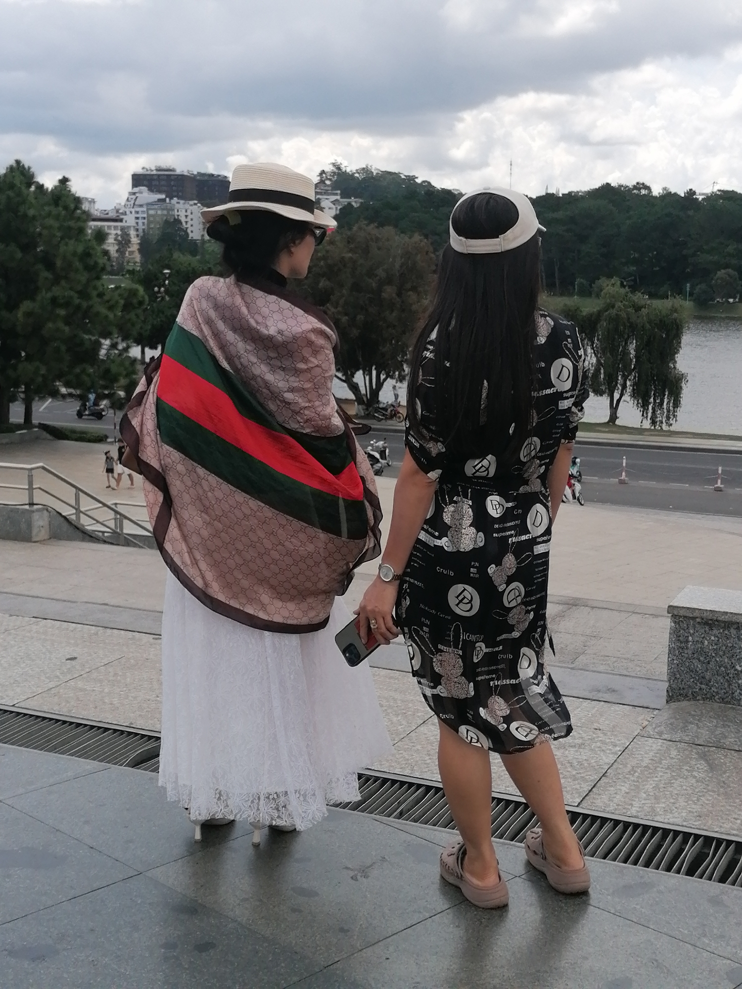 Two women are standing on a raised platform, facing away from the camera. The woman on the left wears a white hat, sunglasses, a white lace dress, and a patterned scarf with green and red stripes. The woman on the right wears a white cap, a black dress with a white pattern, and beige clogs. She is holding a smartphone. In the background, there is a body of water, trees, and buildings, consistent with the landscape around Xuan Huong Lake in Da Lat, Vietnam. The sky is overcast.