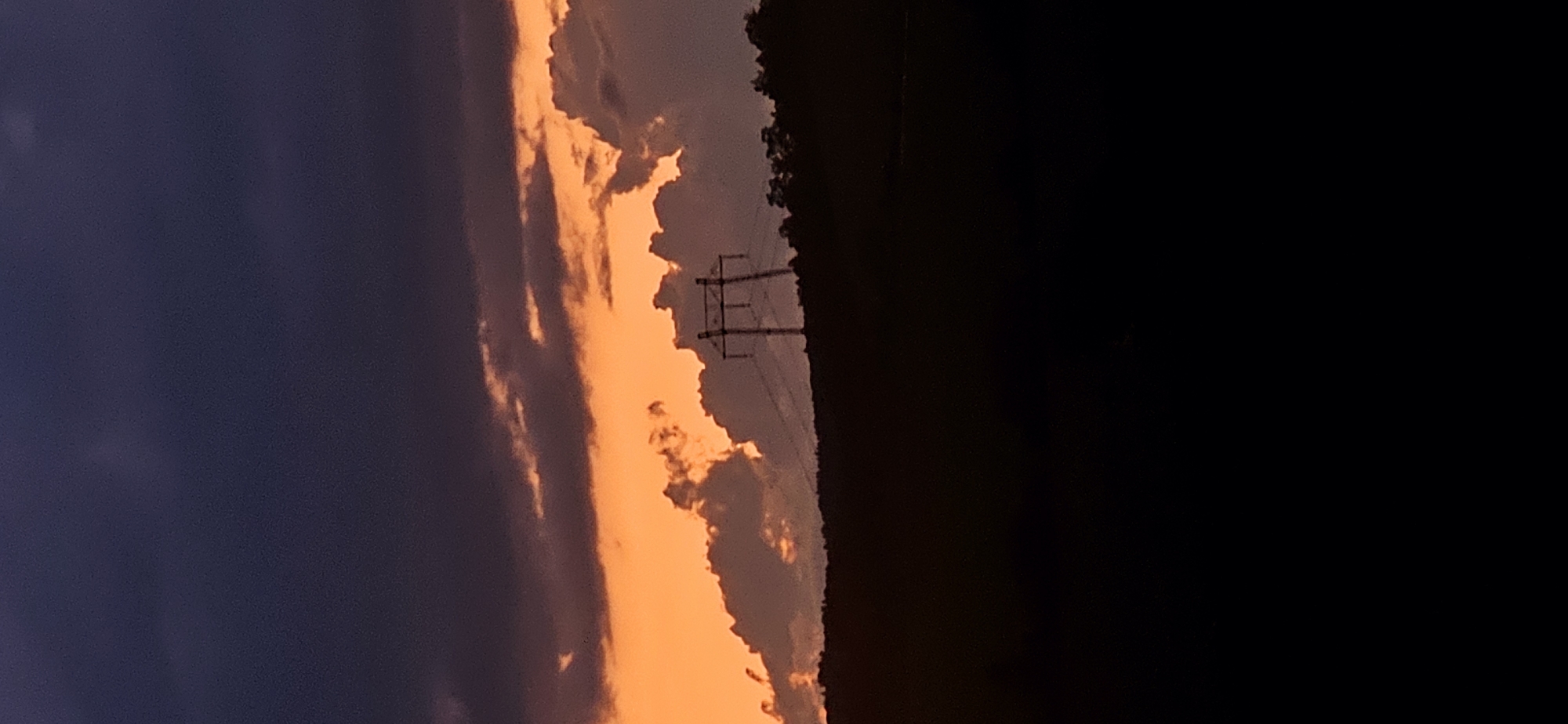 The image depicts an outdoor landscape at dusk or dawn, centered on the area of Gutsal, Bulgaria. The sky is divided into distinct bands: the upper portion is dominated by dark, dense purplish-blue clouds, transitioning to a vivid orange and reddish-orange hue in the mid-section, likely illuminated by a low sun not visible in the frame. Silhouetted against the orange sky, slightly to the right of the vertical center, is a large, multi-lattice steel electrical transmission tower with power lines extending horizontally. Below the bright sky, a dark, undulating horizon line represents a distant treeline or forested hills. The entire lower half to two-thirds of the image, encompassing the foreground and immediate ground beneath the hills, is rendered in deep silhouette as an undifferentiated black expanse. No human figures, animals, or specific actions are discernible.