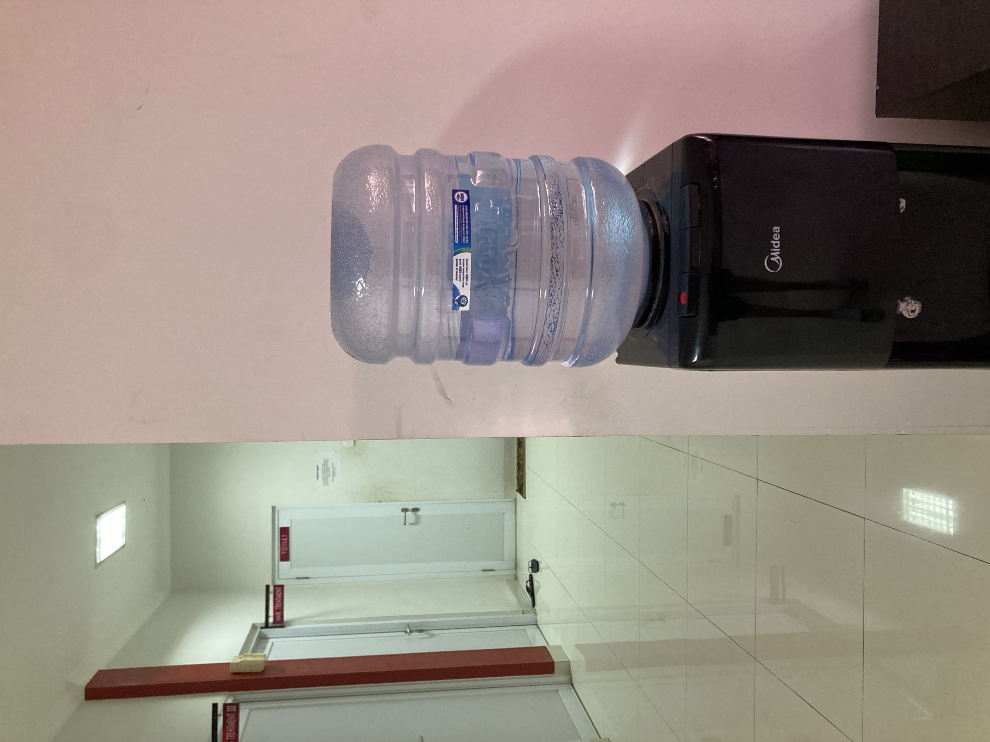 The image shows a hallway in what appears to be a clinic or office building. On the left, there are several doors with signs that read "TREATMENT III," "HAIR TREATMENT," and "TOILET." The hallway floor is tiled and a pair of black flip-flops are on the floor in front of one of the doors. On the right side of the image, there is a black water cooler with a large, clear water bottle on top. The water cooler has the brand name "Midea" on it. The wall behind the water cooler is painted a light pinkish color. The scene is brightly lit by overhead fluorescent lights. The location is not identifiable within the city of Proyonangg, Indonesia, from the provided image.