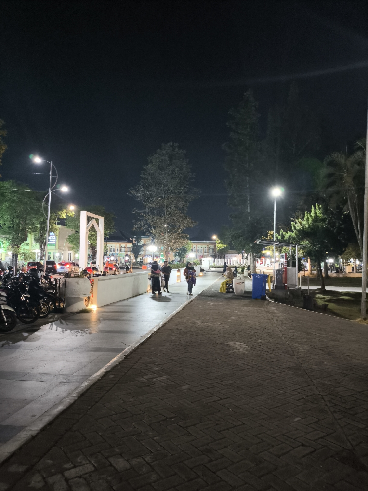 This image depicts an outdoor scene at night, likely in the city of Lumajang, Indonesia, given the context. A paved walkway with a brick pattern occupies the foreground, stretching into the distance. To the left of the walkway, a concrete path is lined with several parked motorcycles. Beyond the concrete path, there is a low, white, wall-like structure with a large, abstract white archway behind it. Several trees are visible throughout the scene, some illuminated by streetlights. In the background, buildings with illuminated windows suggest an urban environment.
Several individuals are present. In the mid-ground, a group of three people are walking away from the camera on the concrete path. Further to the right, another individual is walking towards the camera. A person is also visible sitting on a bench near some trash receptacles. The overall atmosphere is that of a public space at night, possibly a park or a plaza, with artificial lighting illuminating the area. The presence of motorcycles and pedestrian activity suggests it is a place frequented by people in the evening.