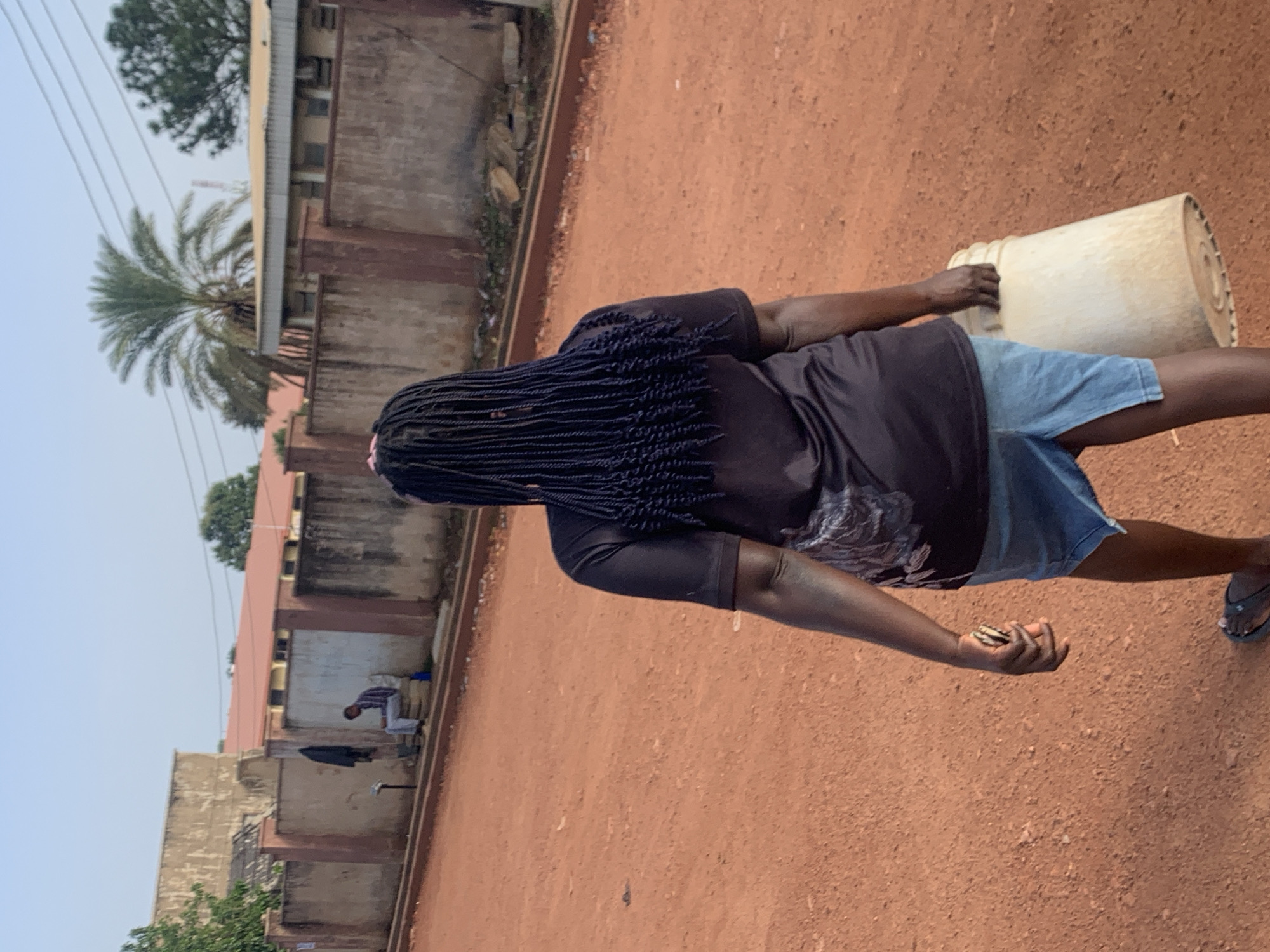 A woman with dark braided hair is walking away from the camera on a reddish-brown dirt road in Jos, Nigeria. She is wearing a dark t-shirt with a faded graphic design and a light blue denim skirt. In her right hand, she carries a large, light-colored plastic bucket. Her left hand is holding a mobile phone. In the background, there is a concrete wall with a reddish-brown cap, and beyond that, buildings with red roofs and some greenery, including a palm tree. To the left, a man in a striped shirt and light pants is seated on a ledge against the wall, with a jacket hanging on a nearby pole. The sky is clear and light blue.