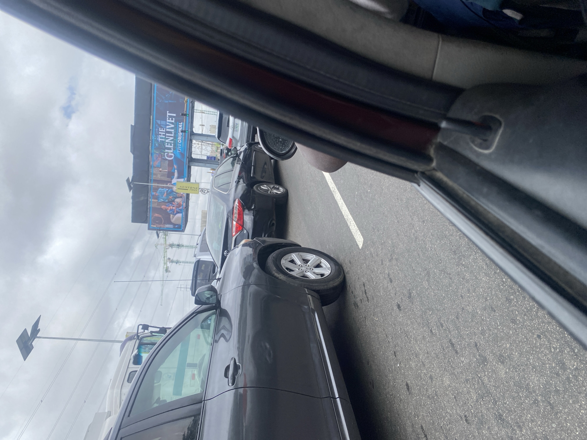 A slightly elevated, medium shot captures a traffic scene in Lekki Phase I, Nigeria, on an overcast day. The perspective is from inside a vehicle, looking out past the driver's side door and window. The foreground is dominated by the dark gray body of a Toyota Highlander SUV, particularly its front left side, tire, and part of the wheel well. The vehicle is stationary in what appears to be a traffic jam on a paved road marked with white lines.
Beyond the SUV, a black Toyota Camry is visible, also stopped. Further in the distance, other vehicles and structures are present. A prominent billboard featuring "THE GLENLIVET LIVE ORIGINAL" is visible above the road, illuminated by daylight. Infrastructure such as streetlights, power lines, and cranes suggest an urban environment under development or maintenance. The sky is cloudy and a light gray, contributing to the diffused lighting of the scene. No people are clearly discernible within the vehicles. The overall impression is one of everyday urban congestion.