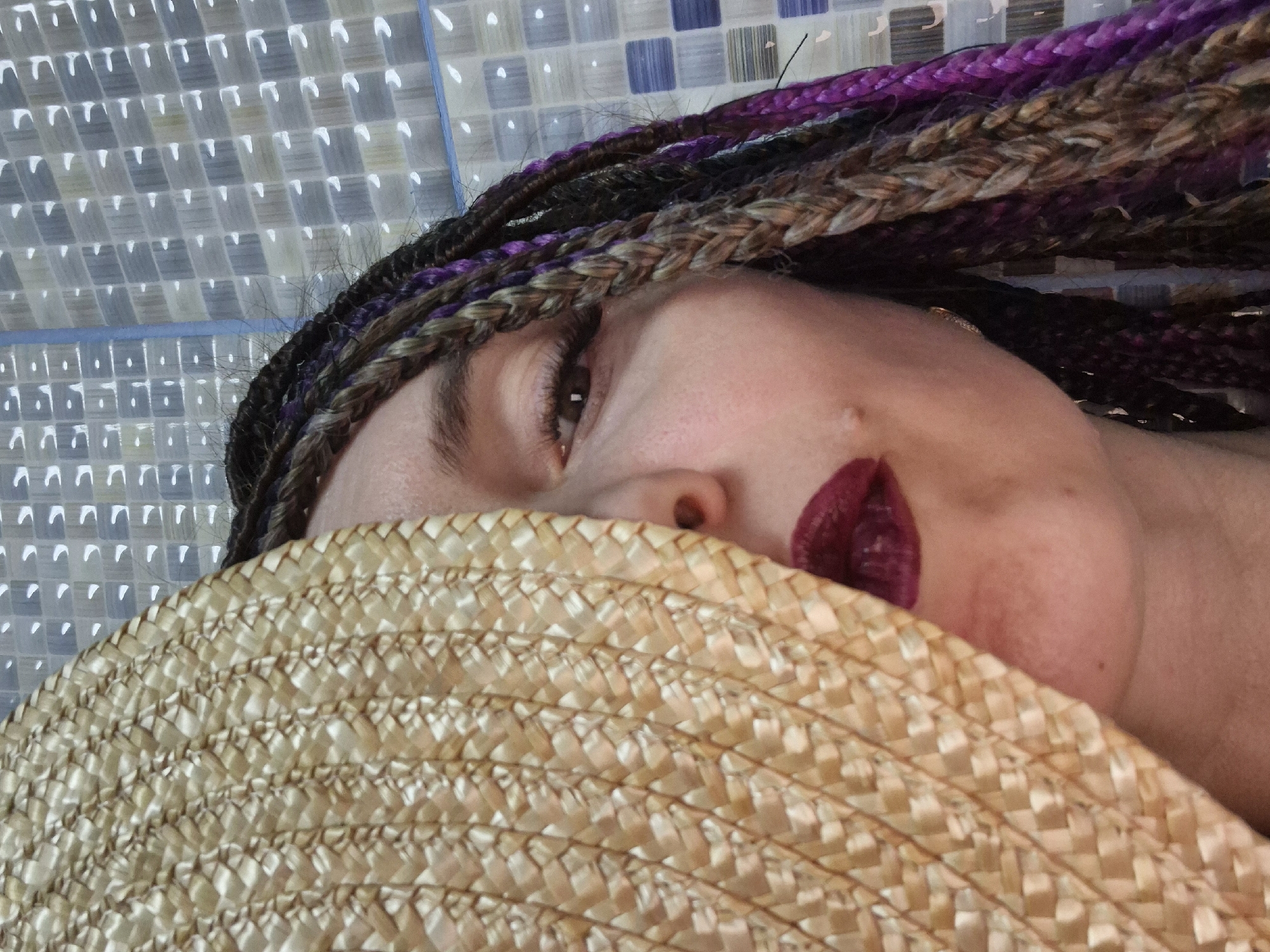 This close-up portrait, oriented diagonally, features a person partially obscured by a straw hat. The visible portion of the face includes the right eye, part of the nose, and the lower right side of the face with lips adorned in dark red or burgundy lipstick. The person's hair consists of multiple braided strands or dreadlocks in various shades of black, brown, blonde, and vibrant purple/magenta, cascading across the upper right portion of the image. The visible eye is dark brown.
A woven straw hat, light beige in color, covers the left side of the person's face, extending across the bottom half of the image. The intricate weave of the hat is clearly visible.
The background is a tiled wall or surface, composed of small, square-shaped tiles in shades of white, light blue, and grey, arranged in a grid pattern. A distinct horizontal blue line, possibly grout or a decorative border, separates different sections or patterns of the tiles in the upper part of the background. The lighting appears even, suggesting an indoor setting. The image captures a casual and intimate moment, likely within a building in Chaikovsky, Russia. No text is visible.