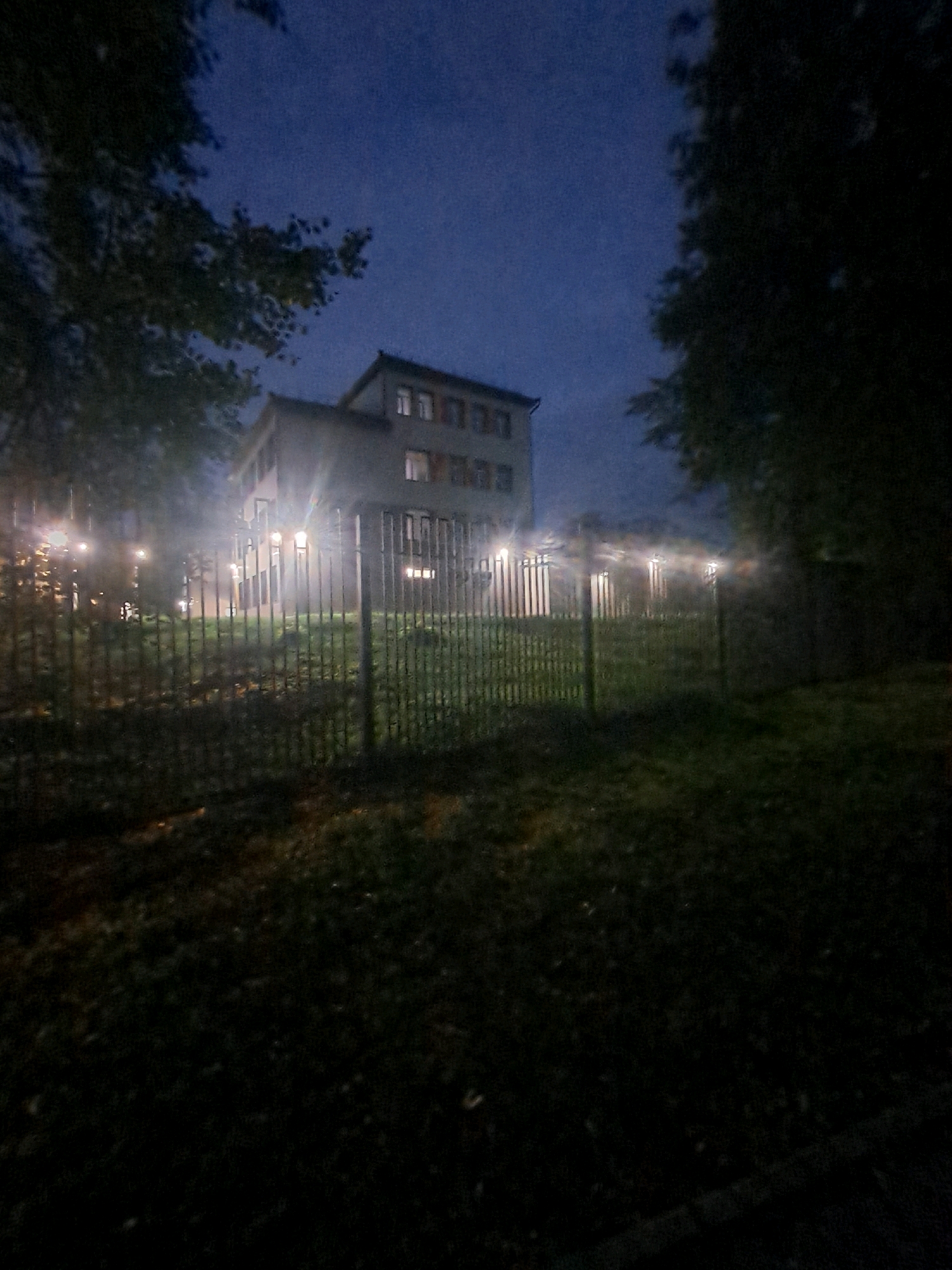 The image depicts a building at night in Elektrostal, Russia. The building is multi-story with windows visible. A fence is in the foreground, and several lights are illuminated along the fence line. Trees are present on either side of the building. The sky is dark blue, suggesting it is nighttime or dusk. The overall impression is one of a residential or institutional building at night, with an atmospheric quality due to the low light and artificial illumination.