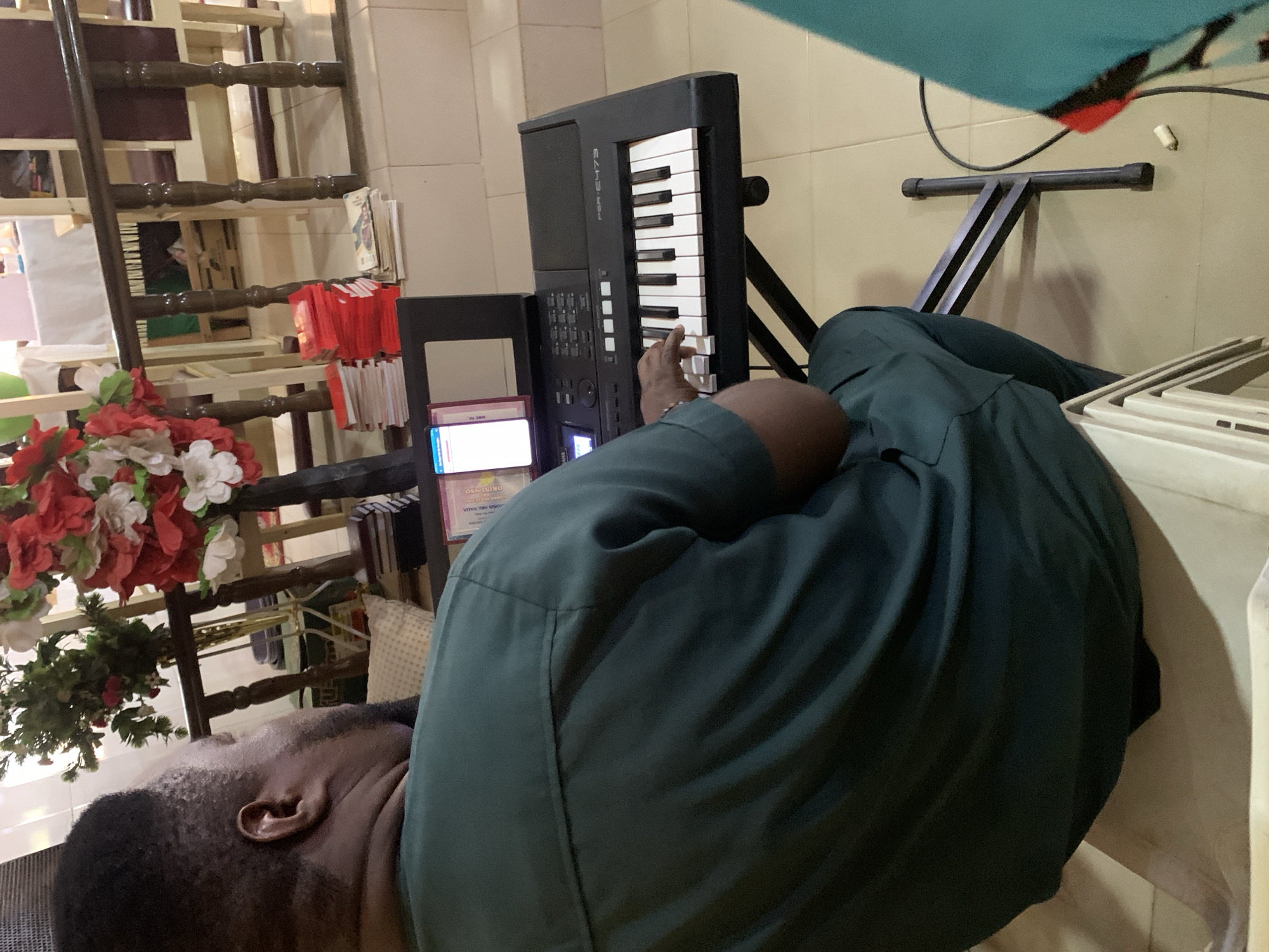 An individual is lying down with their head and shoulders visible in the foreground. They are wearing a dark green shirt. Next to them is a digital piano, with a hand resting on the keys. The piano is on a black stand. A shelf and a ladder are behind the individual and the piano, adorned with red and white decorations and artificial flowers. There is also a stack of papers or leaflets present near the piano. The scene appears to be indoors, with a light-colored wall in the background. Based on the prompt, the location is Akpaka Forest Reser, Nigeria, although the photo does not specifically depict any outdoor elements or characteristics of a forest reserve.