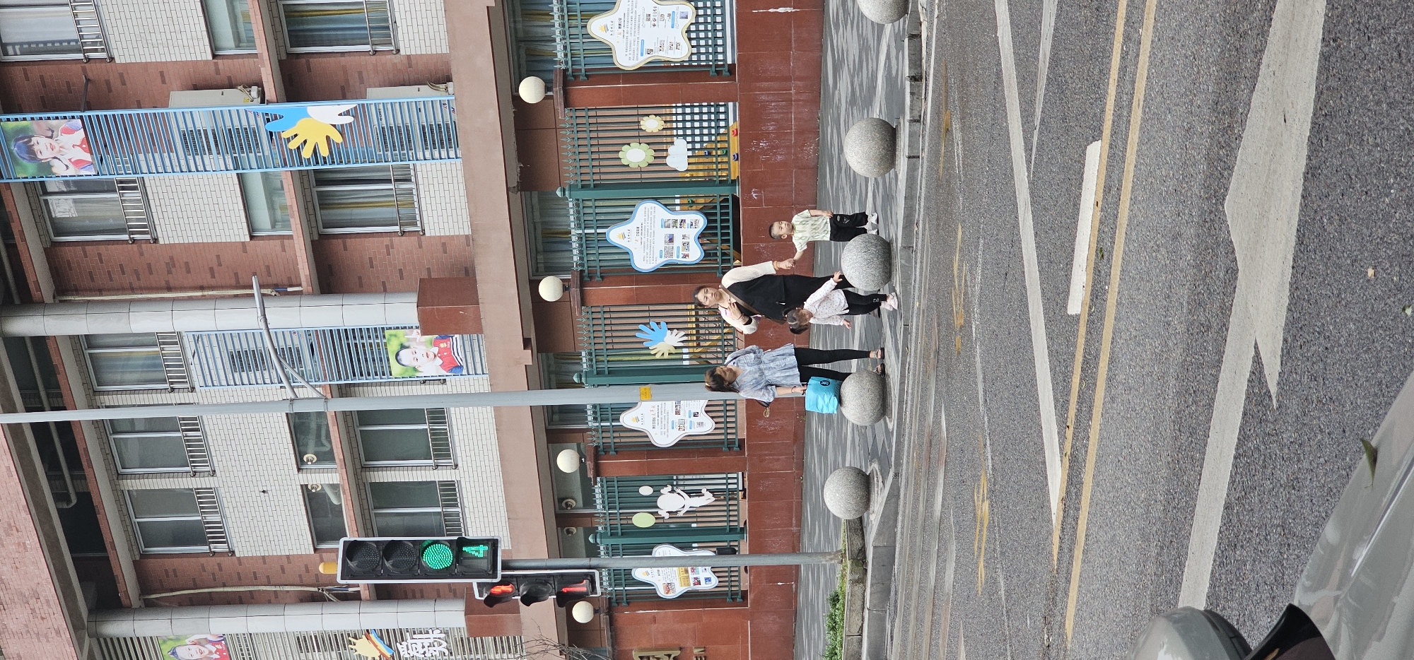 Captured in Jiulongpo District, China, the scene depicts three women and two children standing on a paved area adjacent to a multi-story brick building adorned with educational signage. One woman wears a light blue top and dark pants, carrying a blue bag. Another woman wears a black top and light pants. A small child in light clothing reaches towards the second woman. The building features rows of windows and displays signs with cartoon images. A traffic light with a green signal is visible. The paved area is bordered by a street with painted lanes and a passing vehicle.