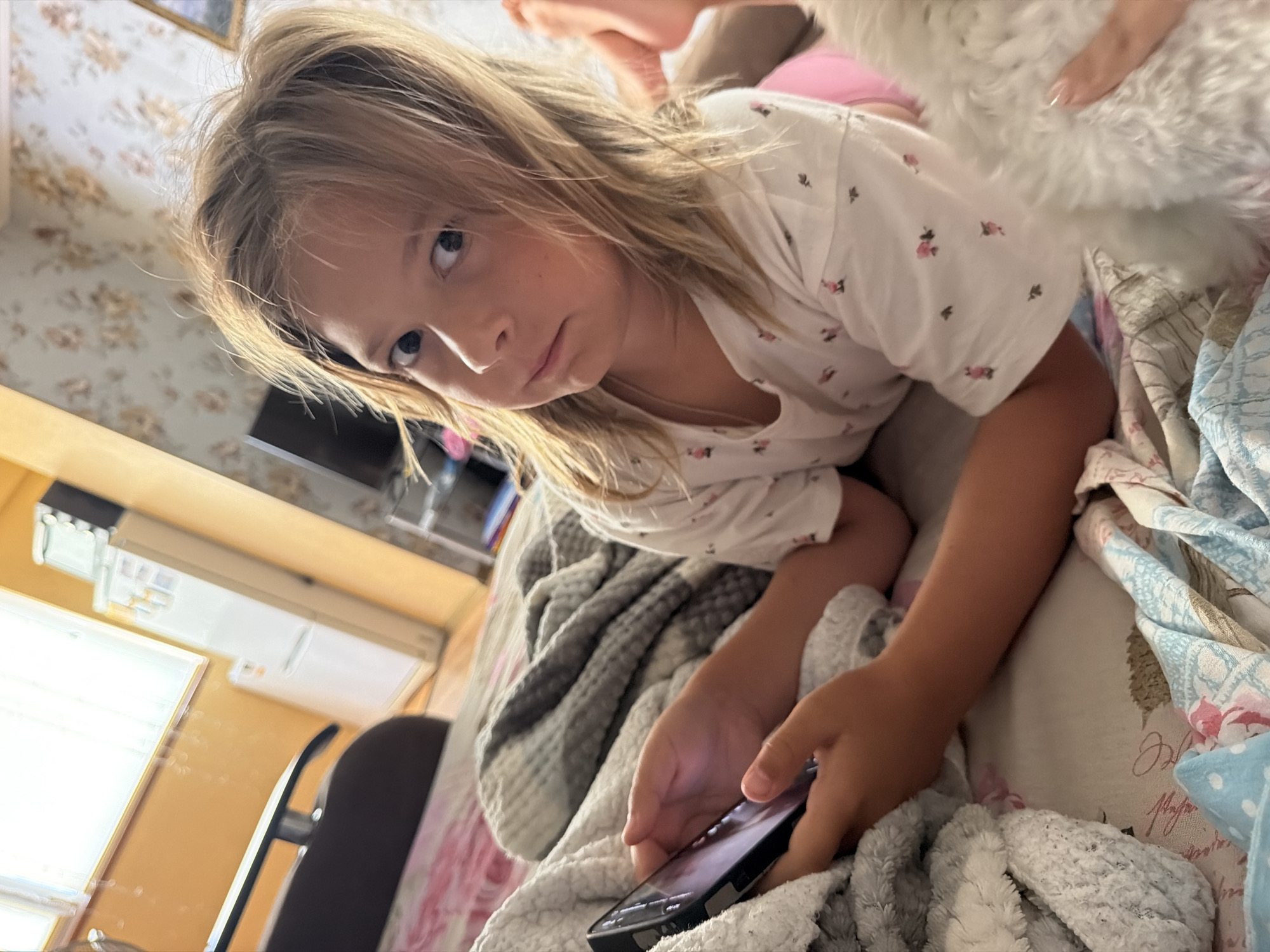 A young girl with blonde hair is lying on a bed, looking at a smartphone. She is wearing a white t-shirt with a floral pattern. The girl appears to be indoors, possibly in a bedroom, with floral wallpaper in the background and a blanket draped over her. The lighting suggests it could be daytime. She is holding the phone in her hands, and her expression is serious or thoughtful. The scene is a close-up shot, focusing on the girl and her activity with the phone, with parts of the bed and surrounding environment blurred.