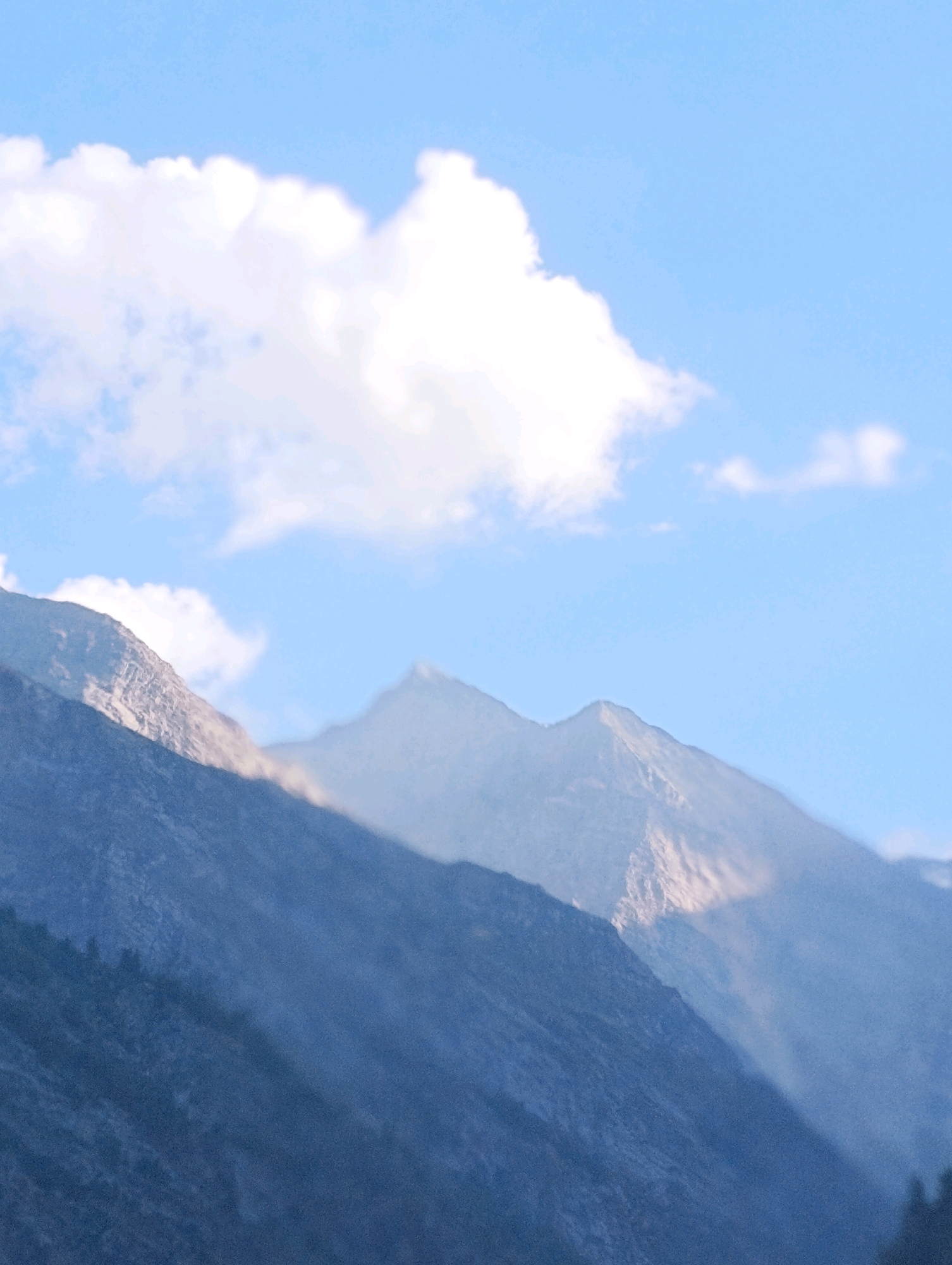 The image captures a mountainous landscape under a clear blue sky with scattered white clouds. In the foreground, a dark, heavily shadowed mountain slope dominates the lower portion of the frame, with hints of vegetation visible on its textured surface. Behind this, the rugged peaks of other mountains rise, some illuminated by sunlight, casting sharp shadows across their faces. The overall atmosphere suggests a high-altitude environment, possibly during the daytime. There are no people, buildings, or any man-made structures visible in this image. The scene is entirely natural, focusing on the geological formations and the sky. The specific location in the city of Gondhla, India, is not visually identifiable within the frame itself, as the image exclusively depicts a natural mountain panorama.
